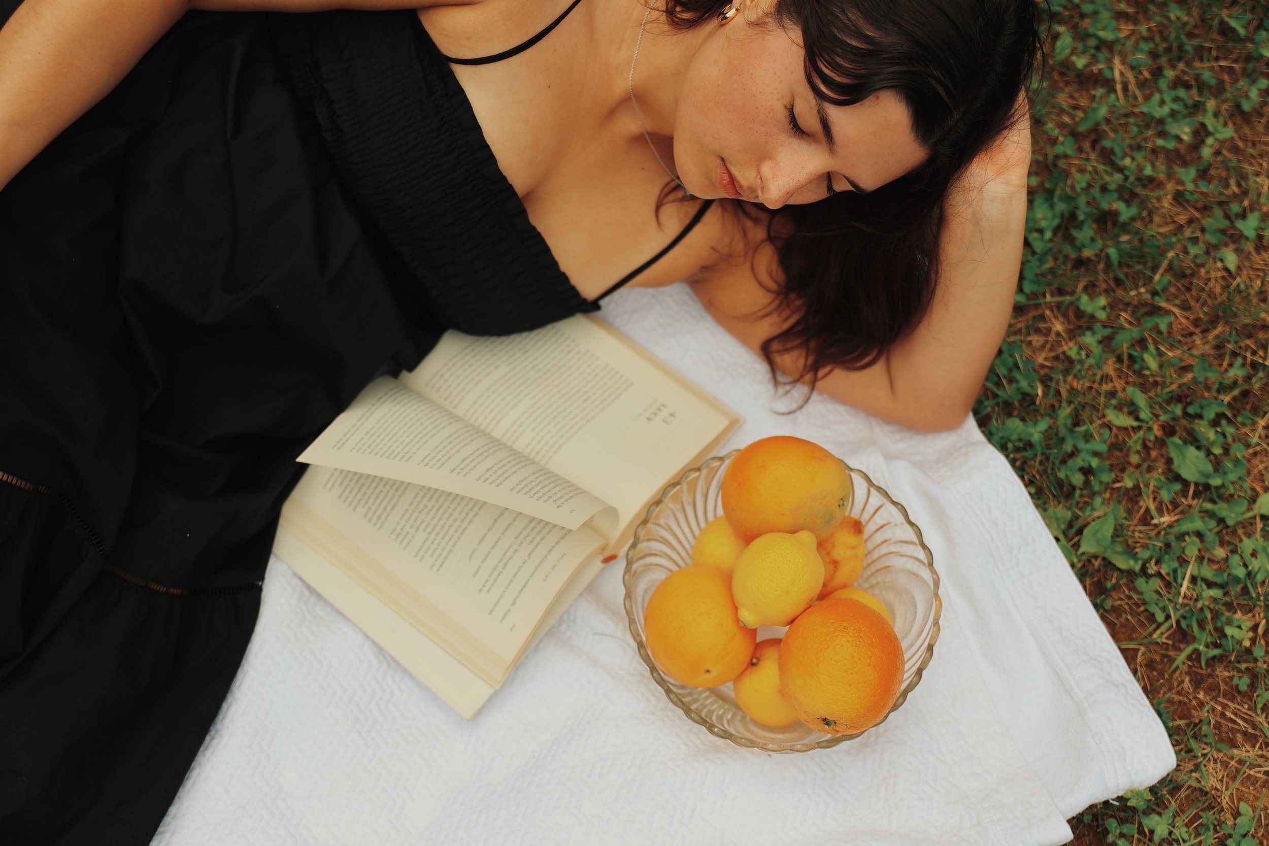 Woman lying on a white blanket outdoors, reading a book, with a bowl of mixed oranges and lemons nearby.