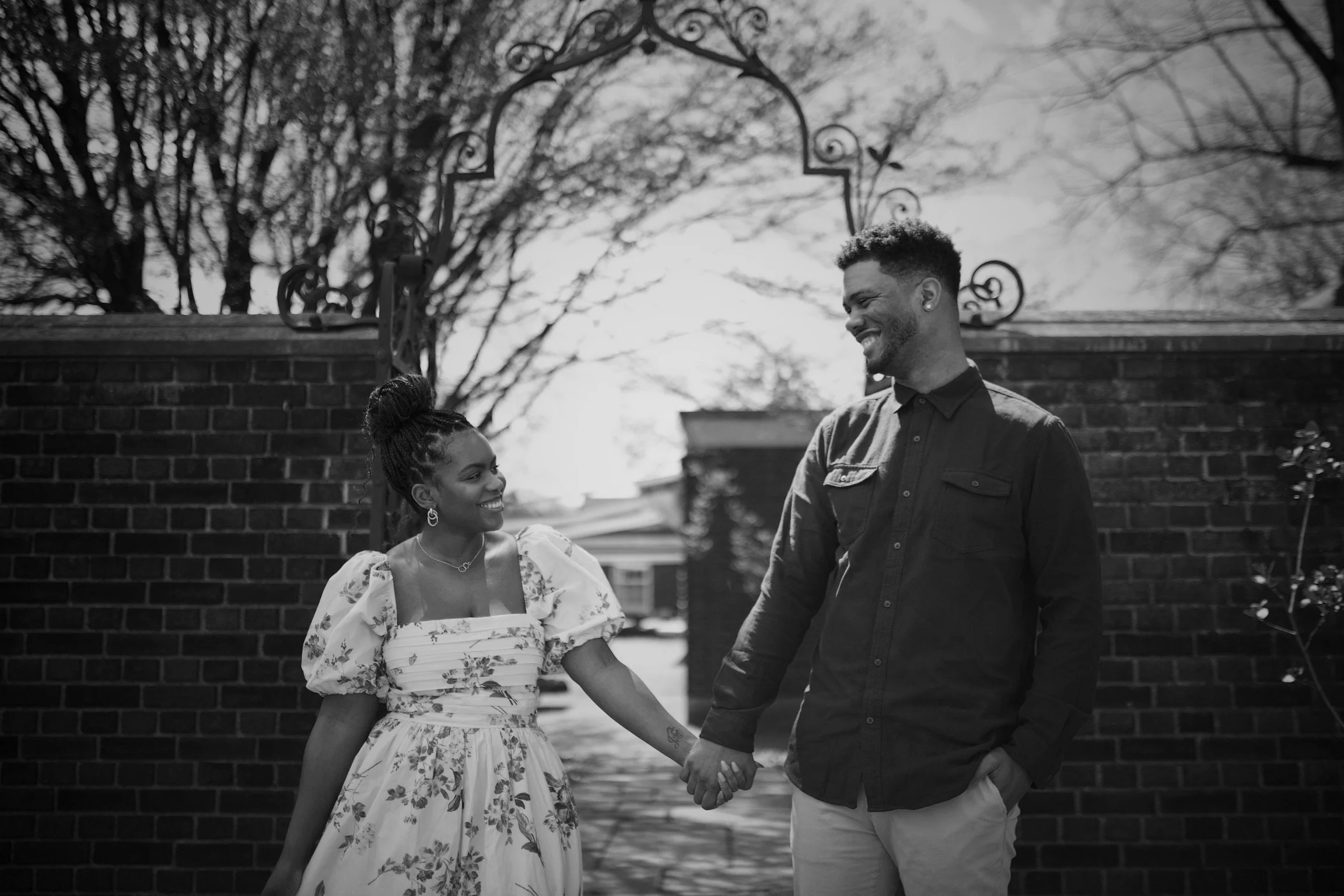 A smiling couple holding hands outdoors, near a brick wall with trees in the background, in black and white.