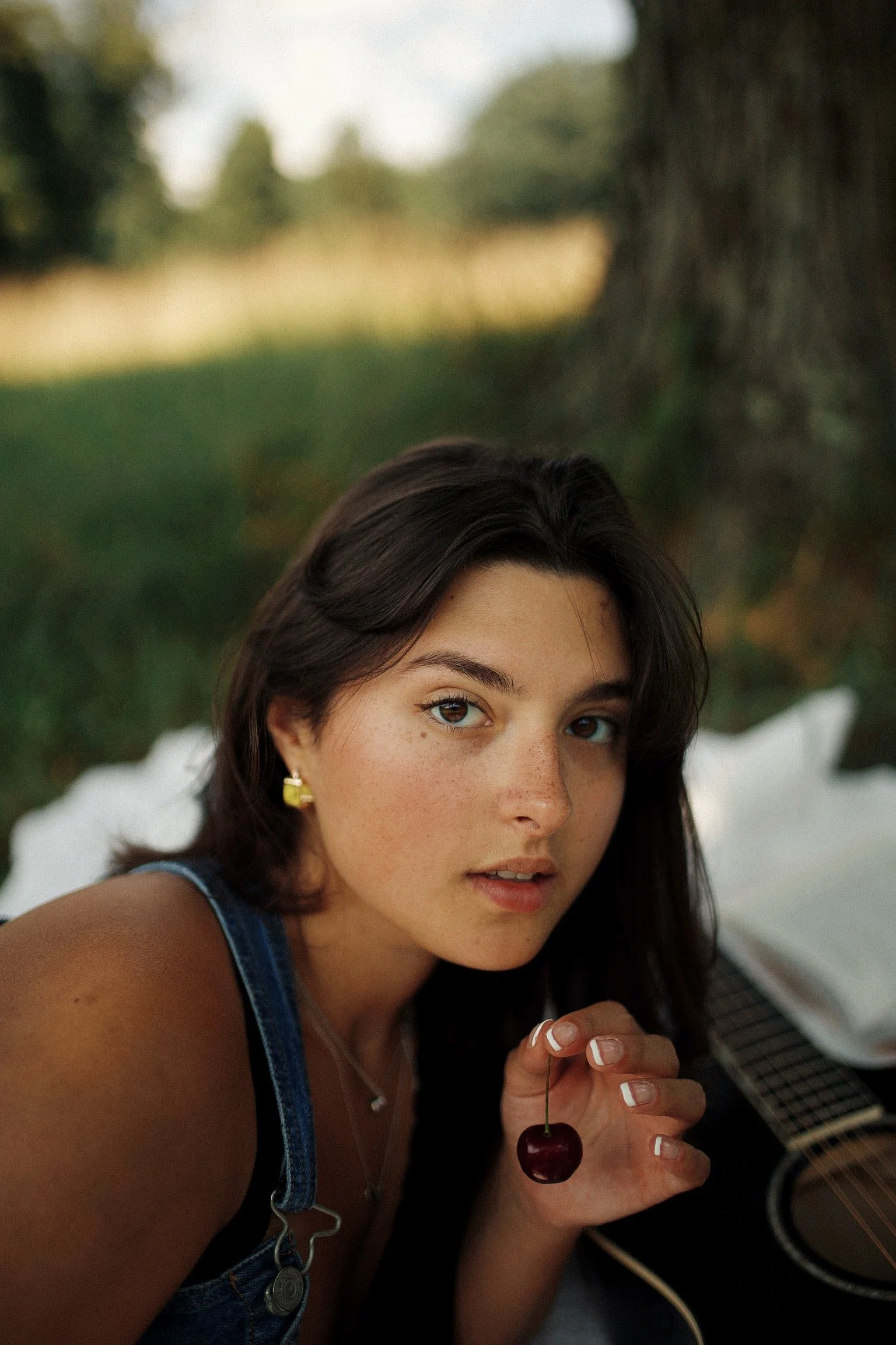 A young woman with dark hair, light skin with freckles, wearing a denim sleeveless top, holding a cherry, sitting outdoors near a large tree with a blurred natural background, and a guitar nearby.
