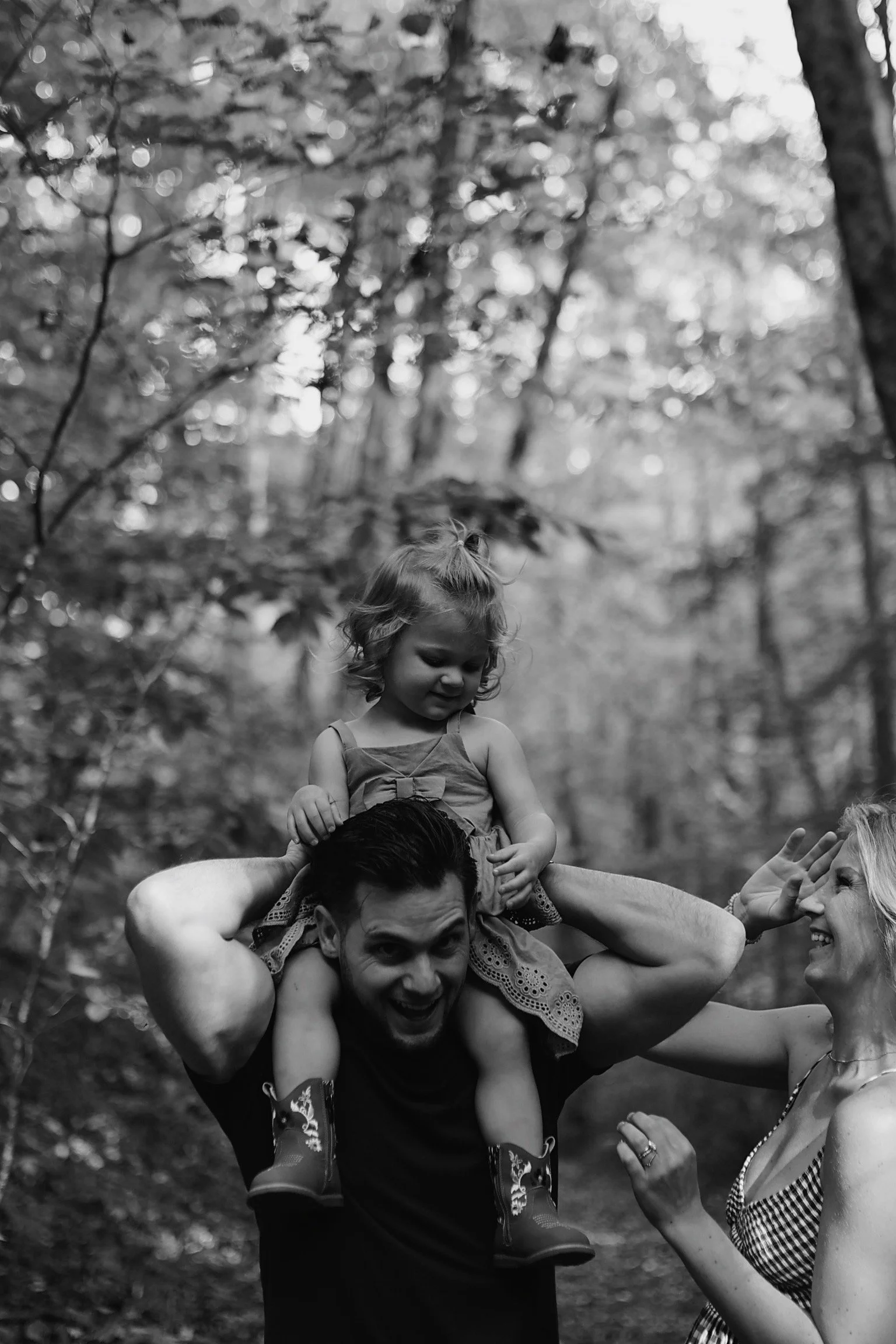 A man carries a young girl on his shoulders while a woman reaches out to her, all smiling in a forest setting.