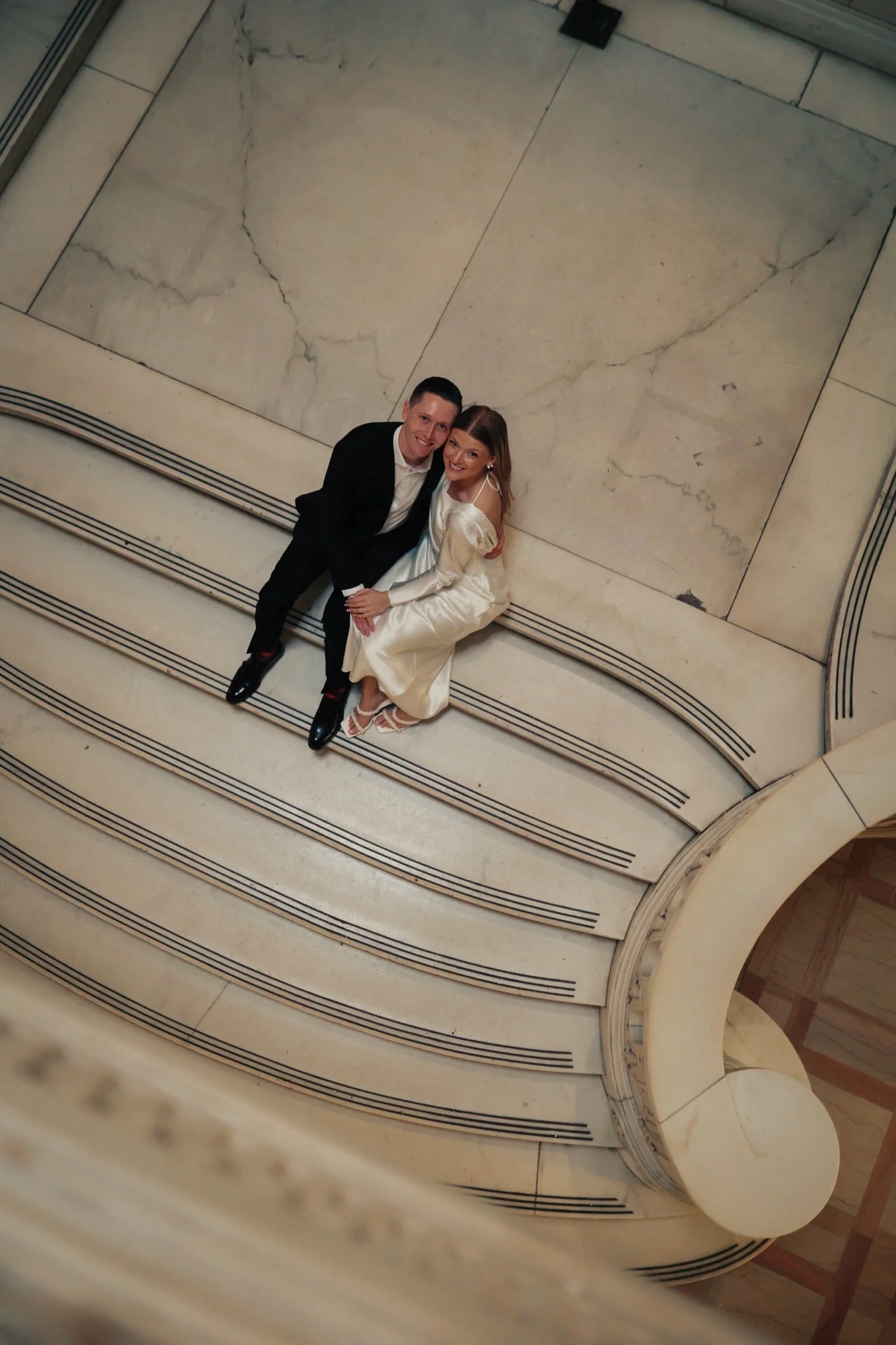 A man and woman sitting on marble stairs, smiling at the camera. The man is wearing a black suit and white shirt, and the woman is in a cream-colored dress with open-toe shoes.