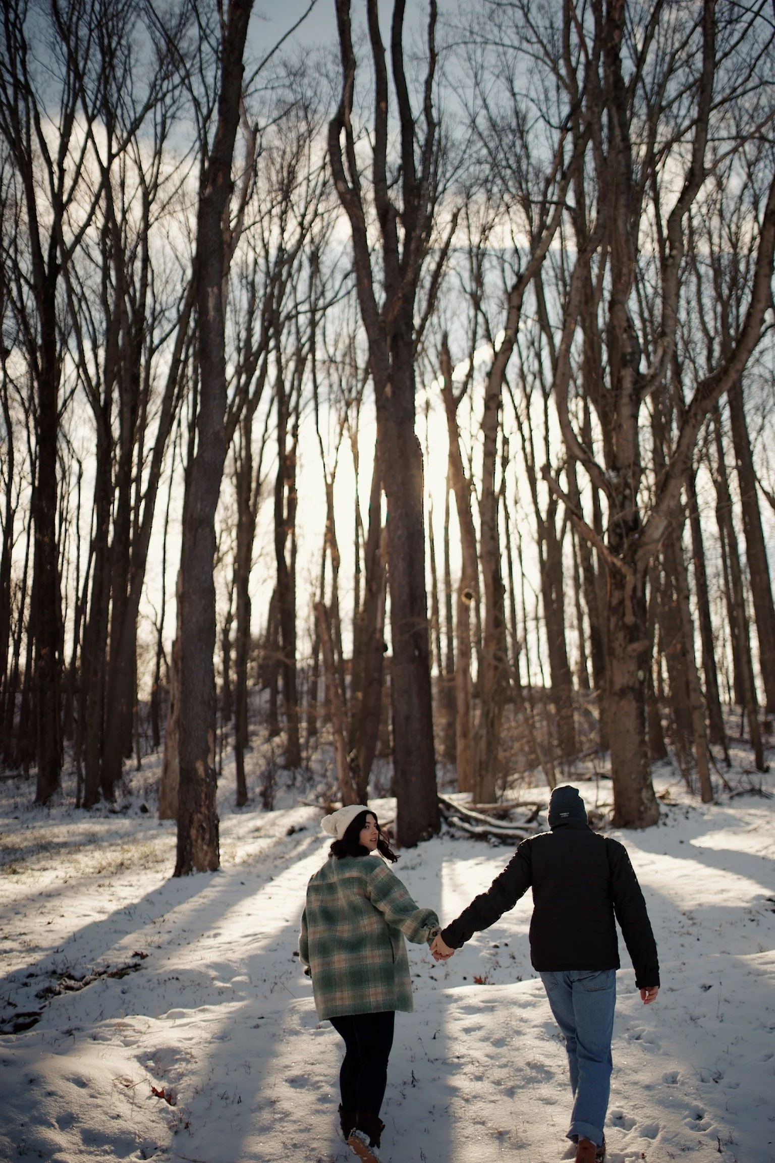 A couple walking hand-in-hand in a snowy forest during late afternoon, wearing winter clothing.