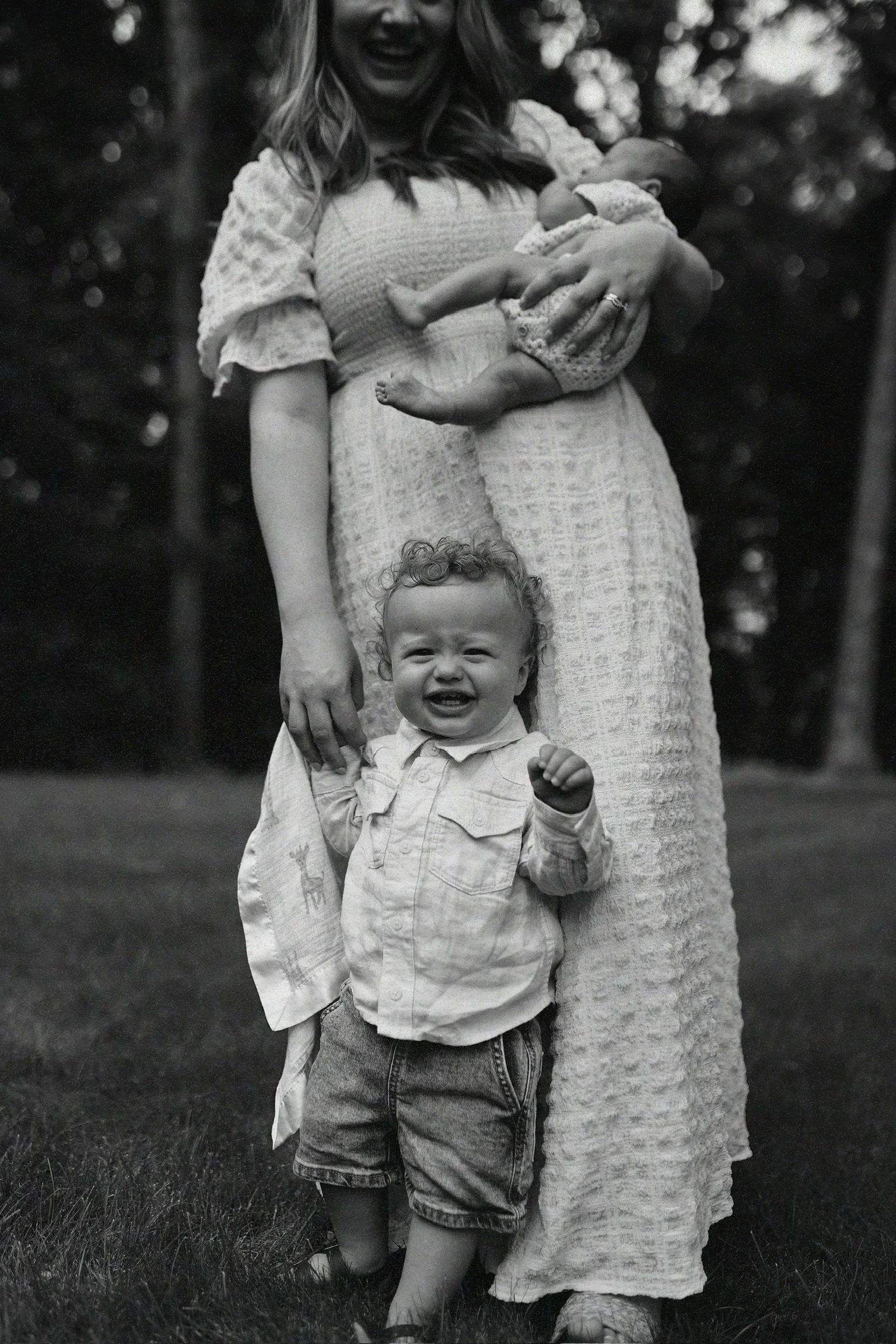 A smiling toddler standing on grass, wearing a long-sleeve shirt and shorts, holding hands with a woman holding a baby in her arms, outdoors in a park or garden.