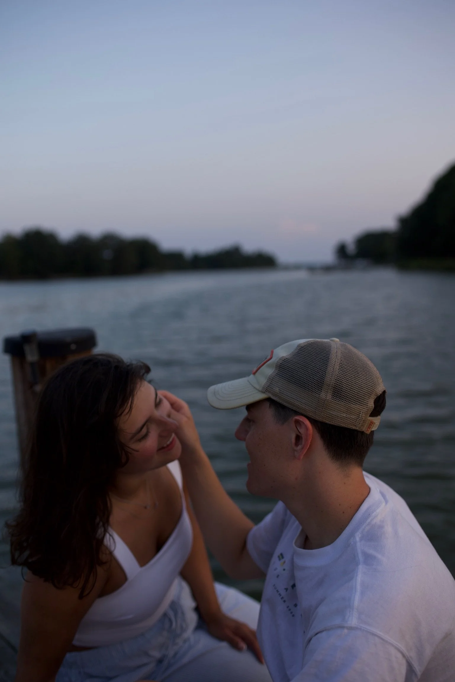 A couple sits closely on a dock by the water during dusk, smiling and touching faces, with a river and trees in the background.