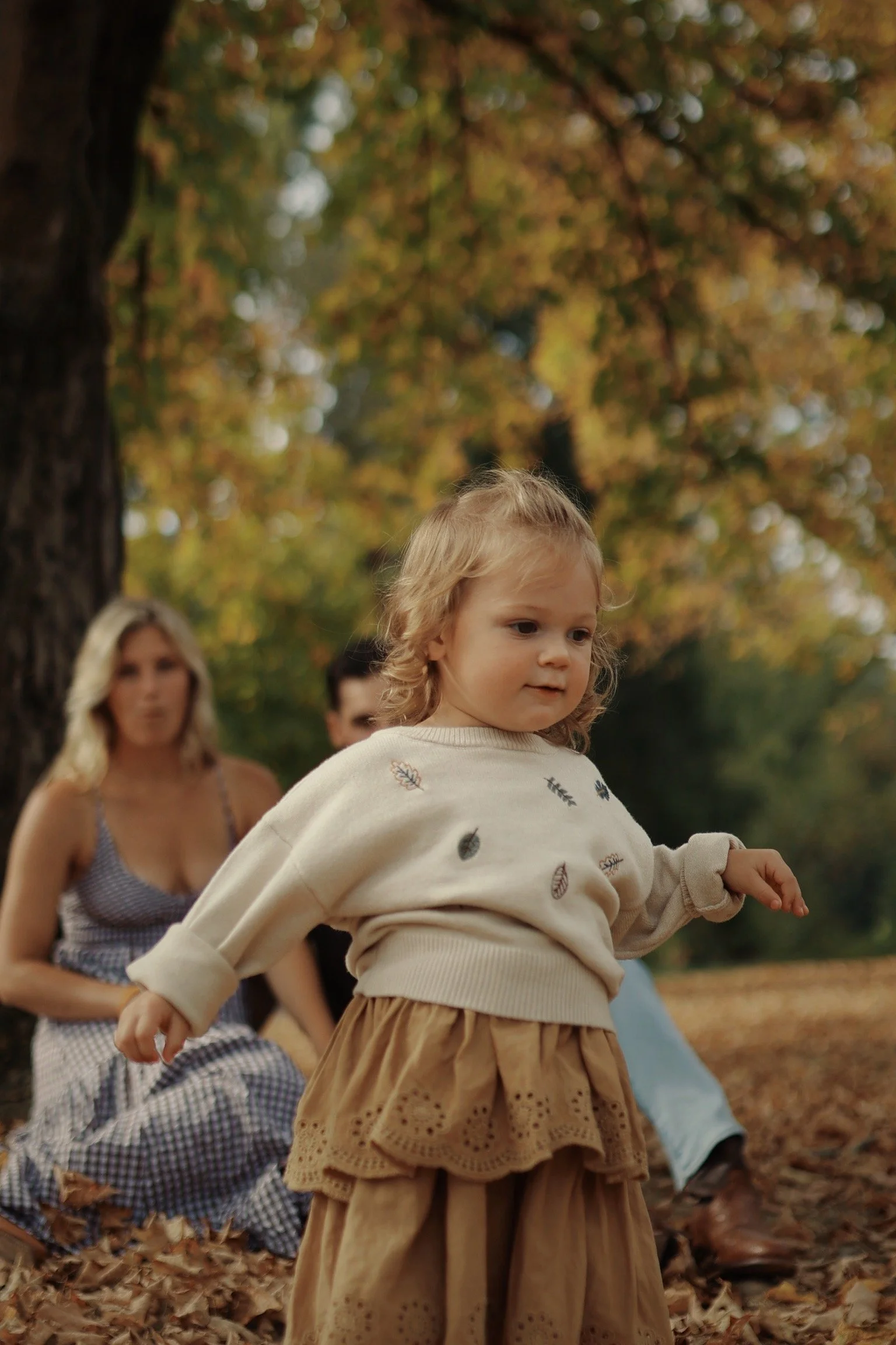 A young girl with blonde curly hair standing outdoors in fall, wearing a cream sweater with leaf patterns and a tan skirt. Two women are sitting behind her among fallen leaves, with autumn trees in the background.