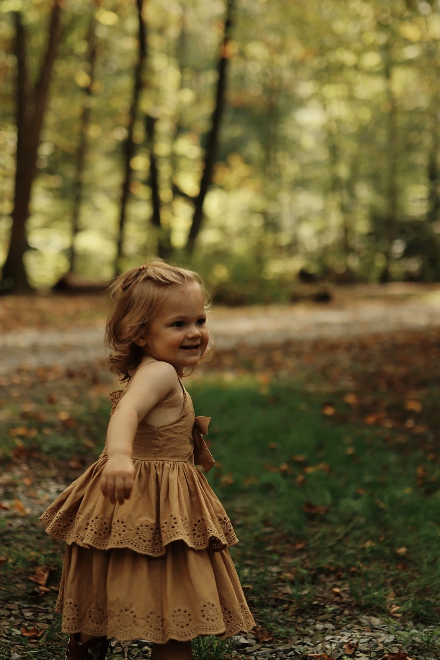 A young girl with blonde hair wearing a tan dress with eyelet lace details is smiling and playing outdoors in a wooded area with fall foliage.