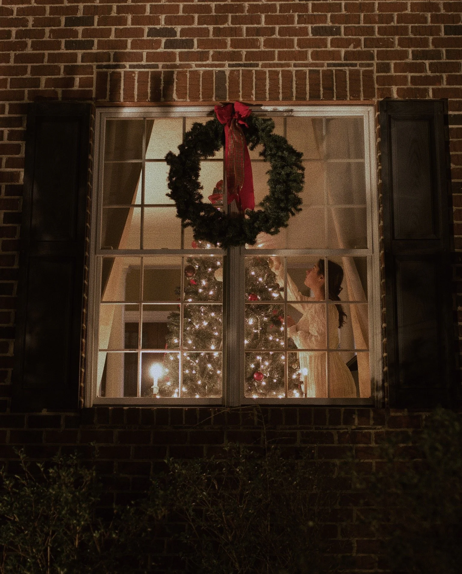 A view through a window showing a decorated Christmas tree with lights and ornaments inside a house, with a young girl in a light-colored dress looking at the tree, and a wreath with a red bow hanging above the window.