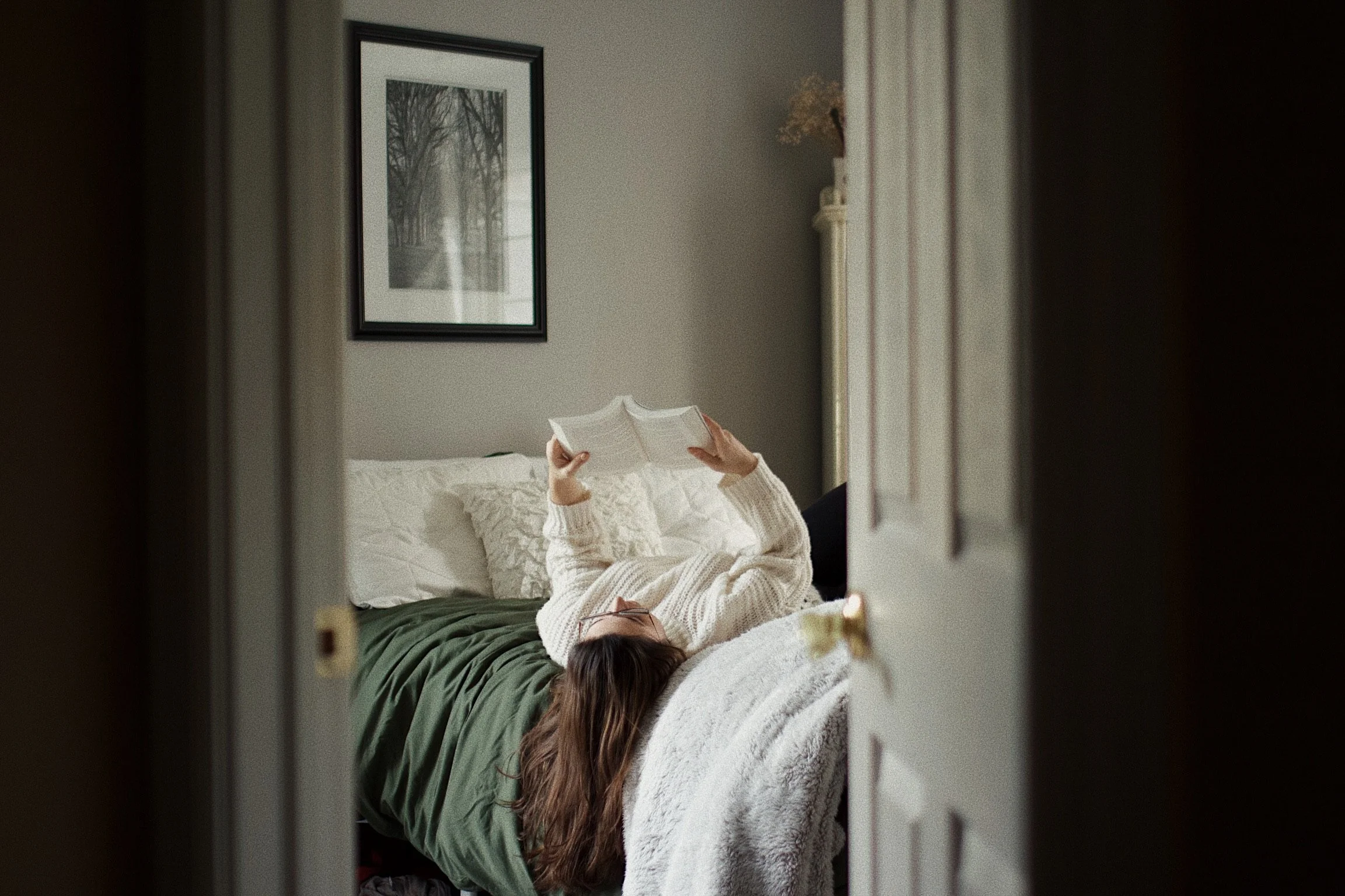 Person lying on a bed reading a book, viewed through a partially open door. The bed has a green blanket and white pillows. A framed black and white photograph of trees hangs on the wall above the bed, and a white dresser with a potted plant is visibl