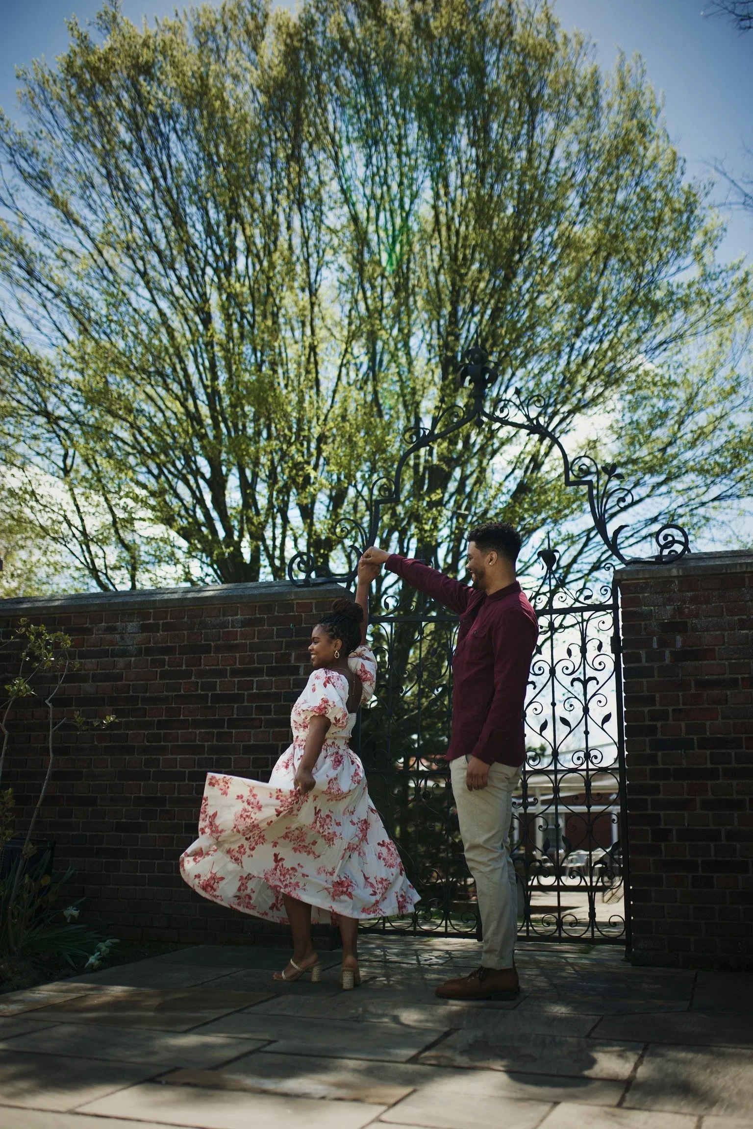 A couple dancing outdoors near a decorative wrought iron gate with a large green tree and blue sky in the background.