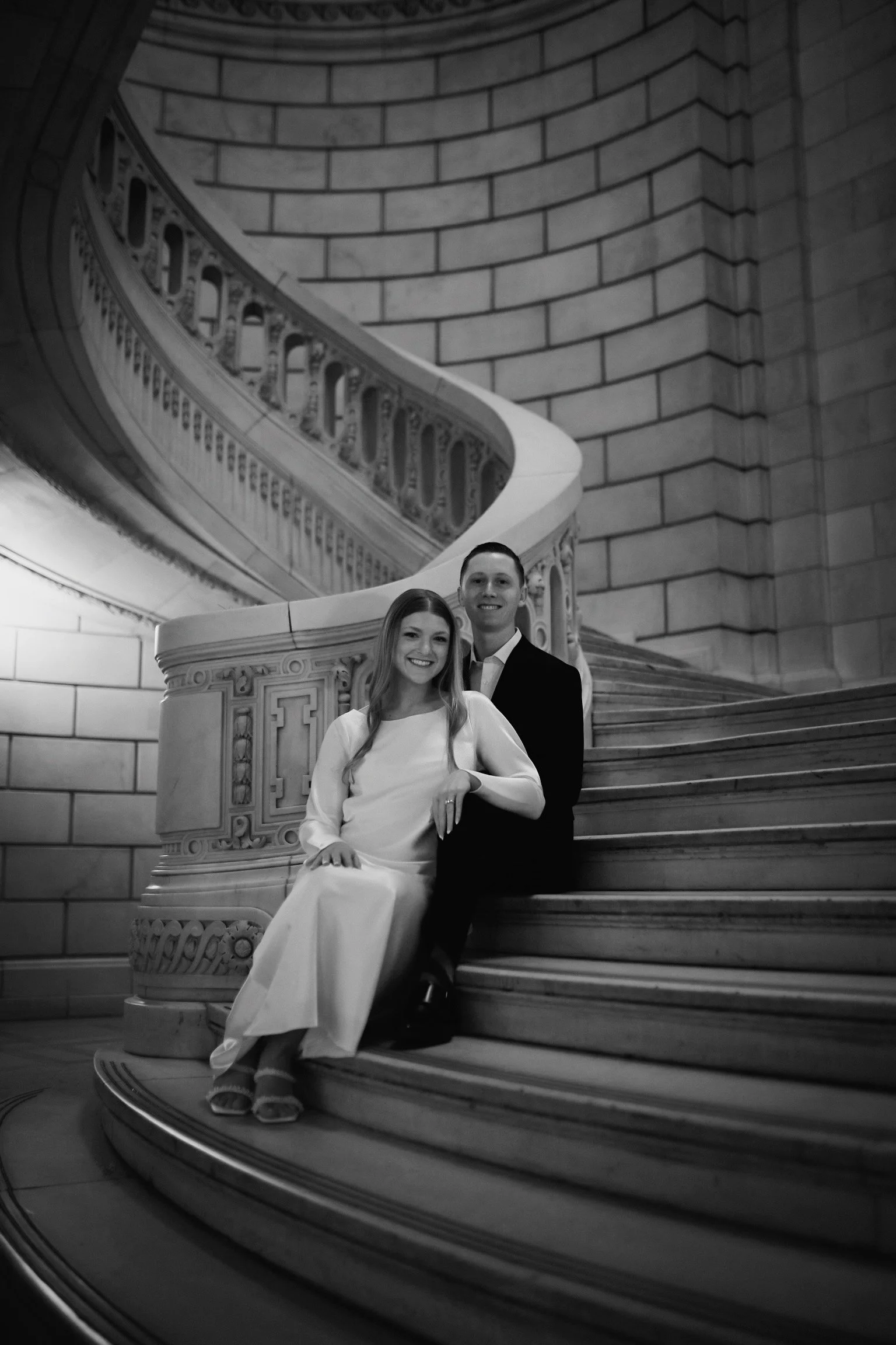 A black and white photograph of a smiling couple sitting on grand staircase with ornate railing, inside large building with high walls and archways.