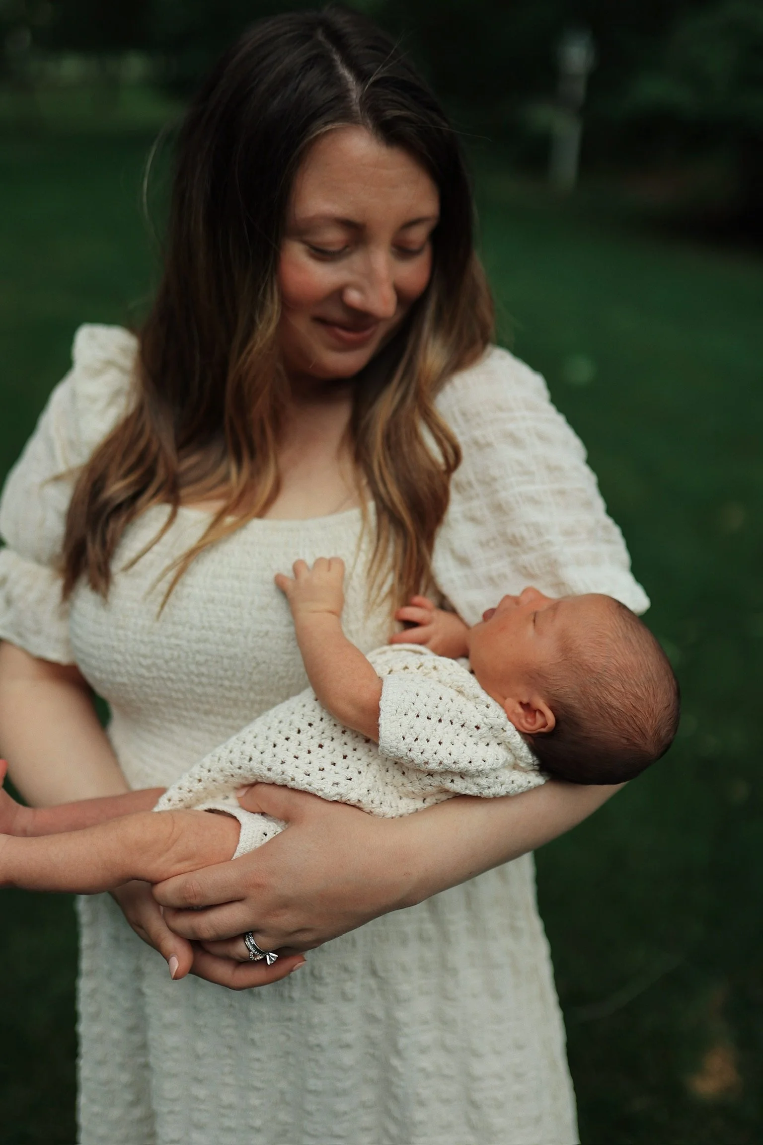 A woman in a cream-colored dress holding a newborn baby in a matching knit outfit, outdoors in a green area.