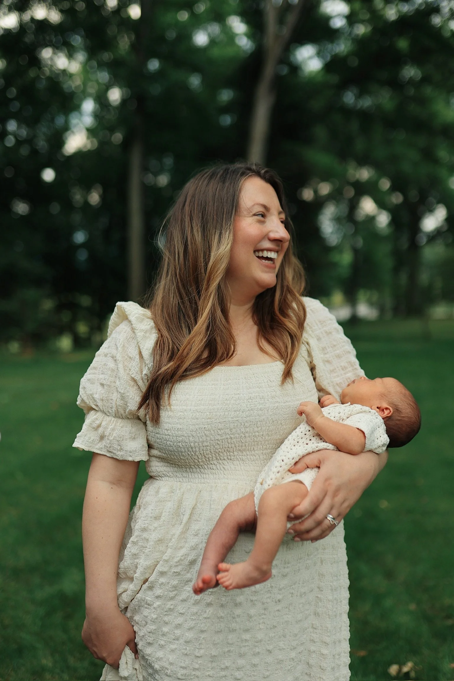 A woman holding a baby outdoors in a park, smiling and looking to the side.
