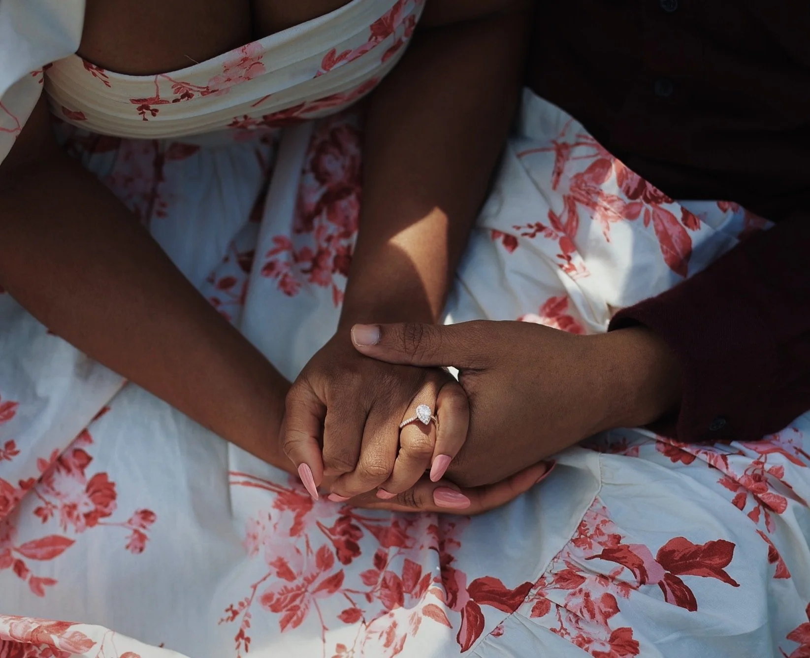 Close-up of two people holding hands, one with a large diamond engagement ring, lying on a floral-printed white sheet. One person's hand is darker and the other's hand is lighter.