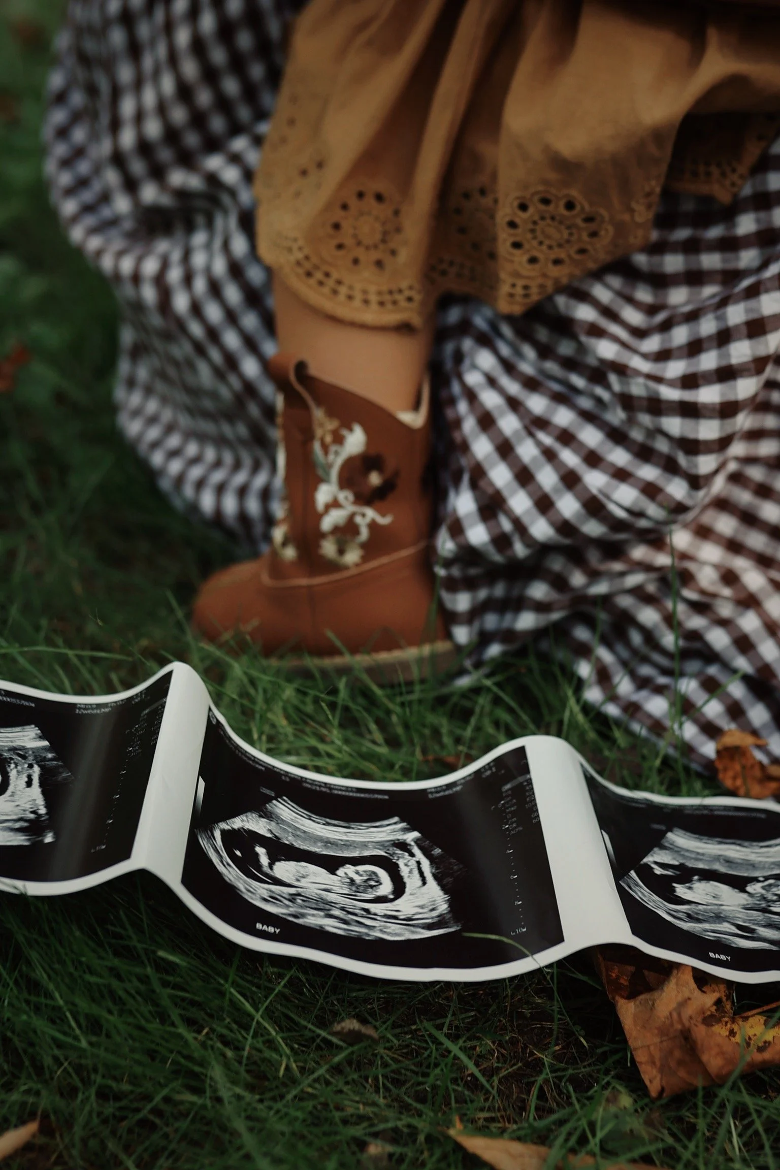 A person sitting on grass holding an ultrasound image of a baby. The person is wearing a brown checkered dress, a beige shawl, and brown embroidered boots.