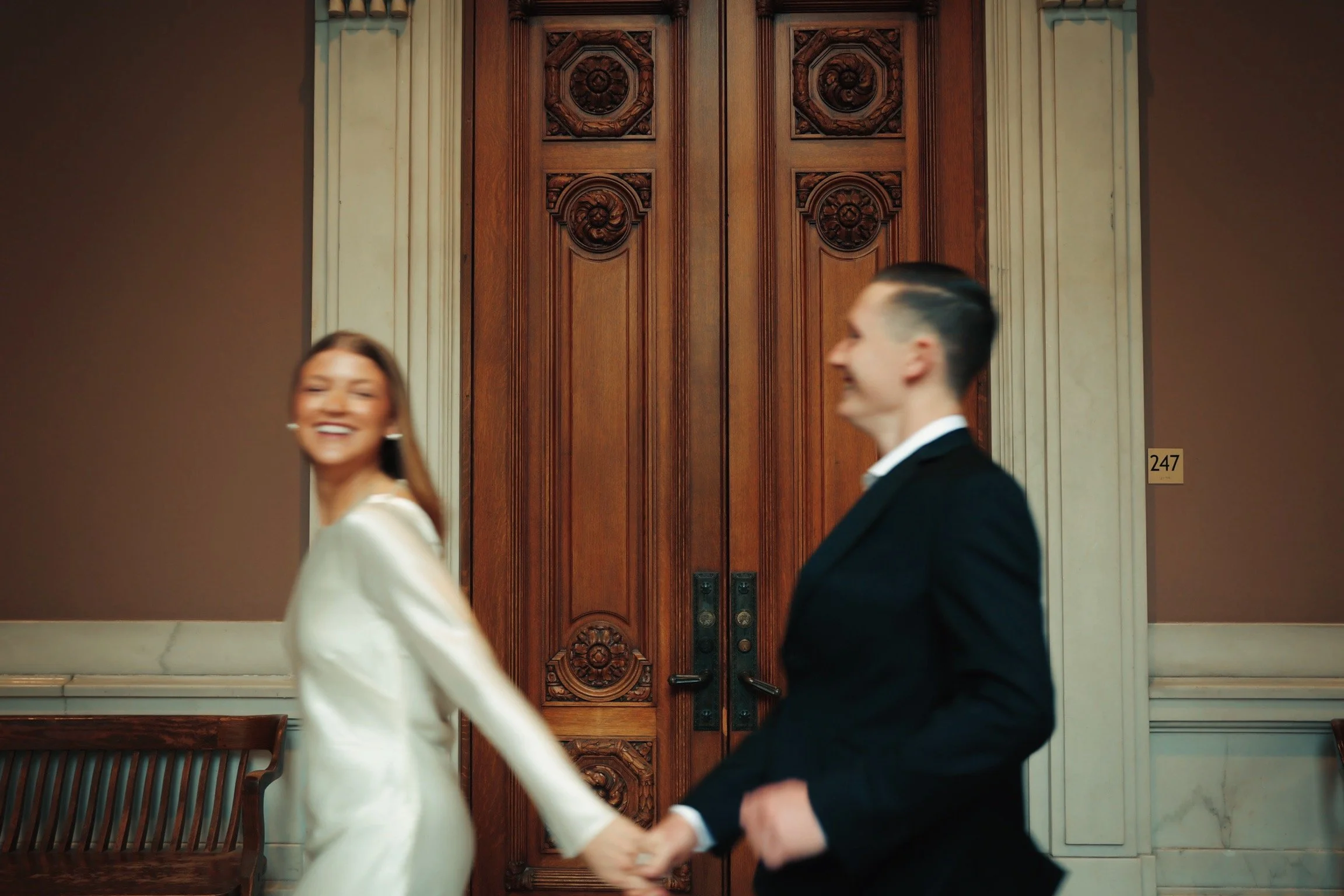 A joyful couple holding hands, smiling and walking in front of a large wooden door in an elegant, classic interior with benches and decorative wall features.