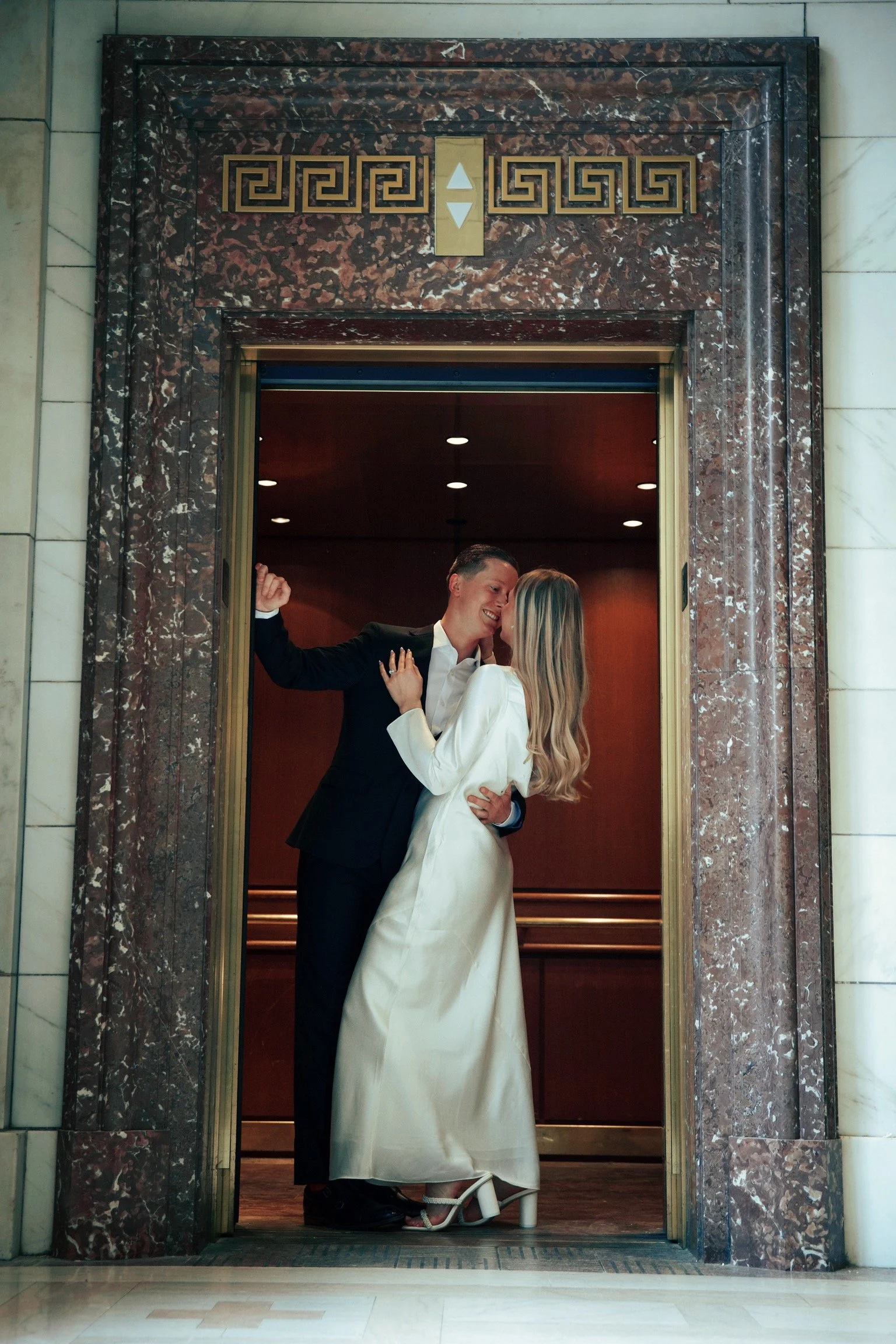 A couple sharing a kiss and dancing in an elevator, with the woman wearing a white dress and the man in a black suit.