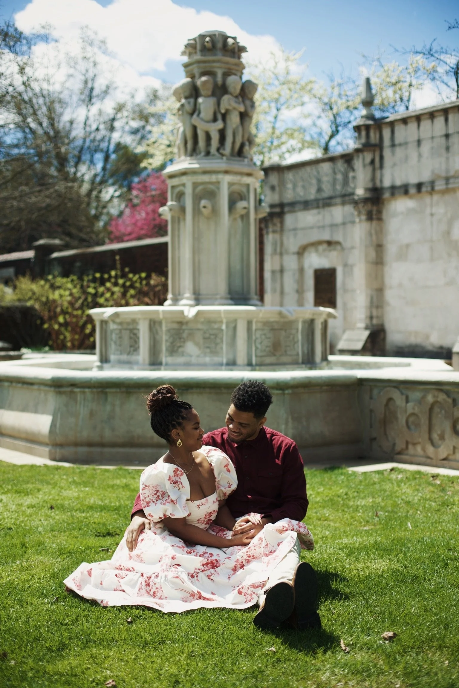 A couple sitting on the grass in front of a marble fountain in a garden, smiling at each other. The woman is wearing a white dress with red floral patterns, and the man is dressed in a dark red shirt.