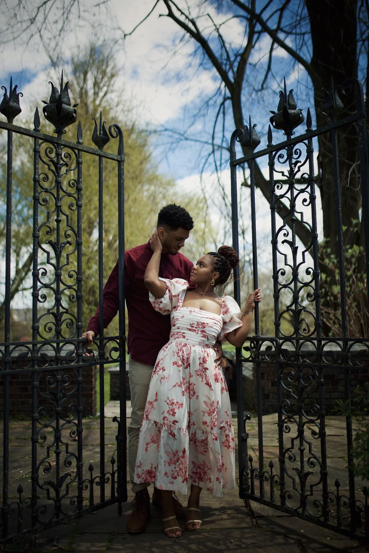 A young couple stands in an open wrought iron gate with intricate designs, surrounded by trees and a partly cloudy sky. The woman, dressed in a floral off-shoulder dress, looks up at the man, who wears a dark red shirt and light-colored pants. She to
