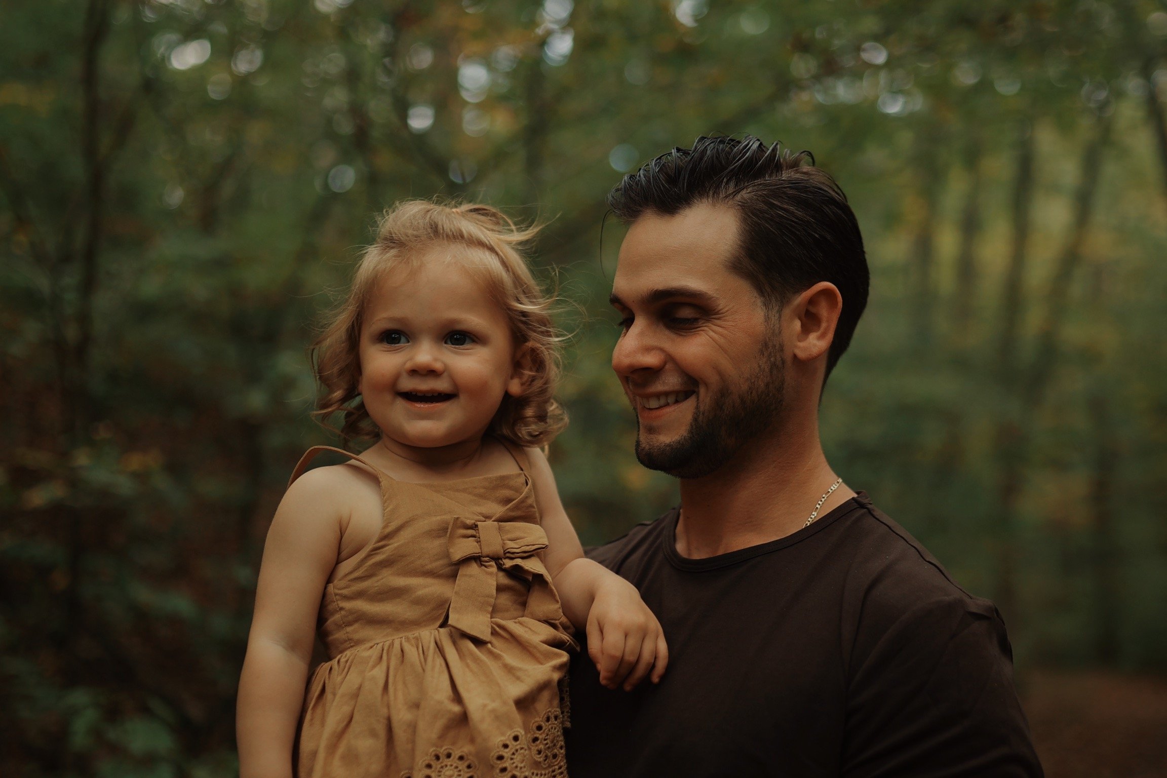 Man holding a young girl outdoors in a wooded area, both smiling and enjoying the moment.