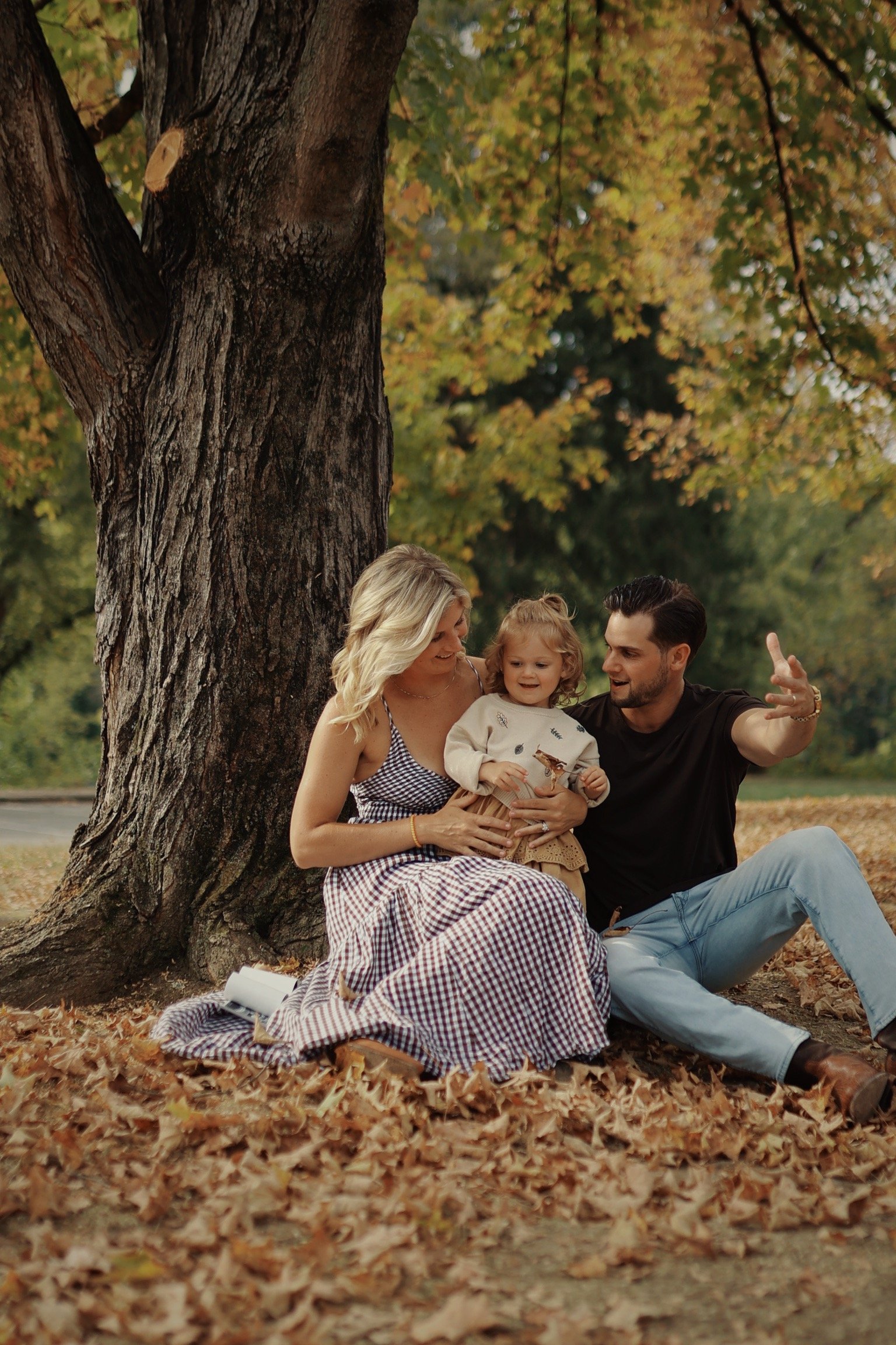 A family of three enjoying a fall day under a large tree, sitting on leaves, with the mother holding a child and the father gesturing while talking.