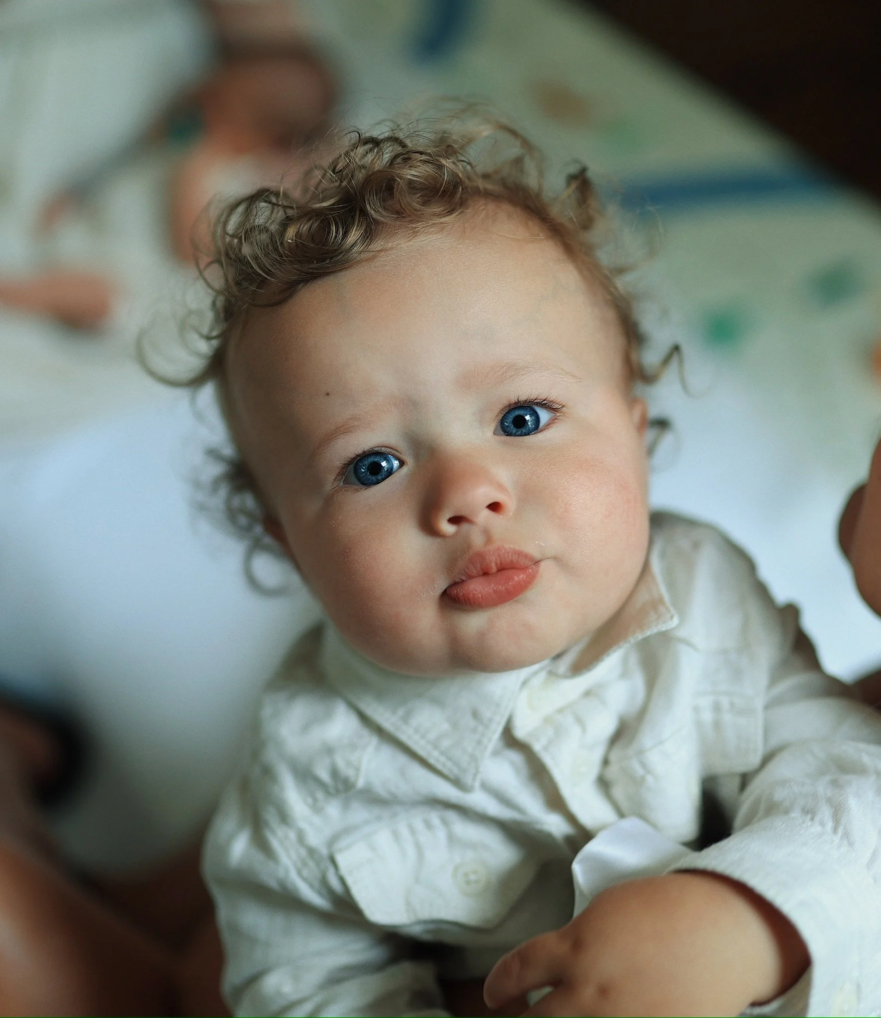 Close-up of a young child with curly blonde hair and blue eyes, wearing a white shirt, looking at the camera.