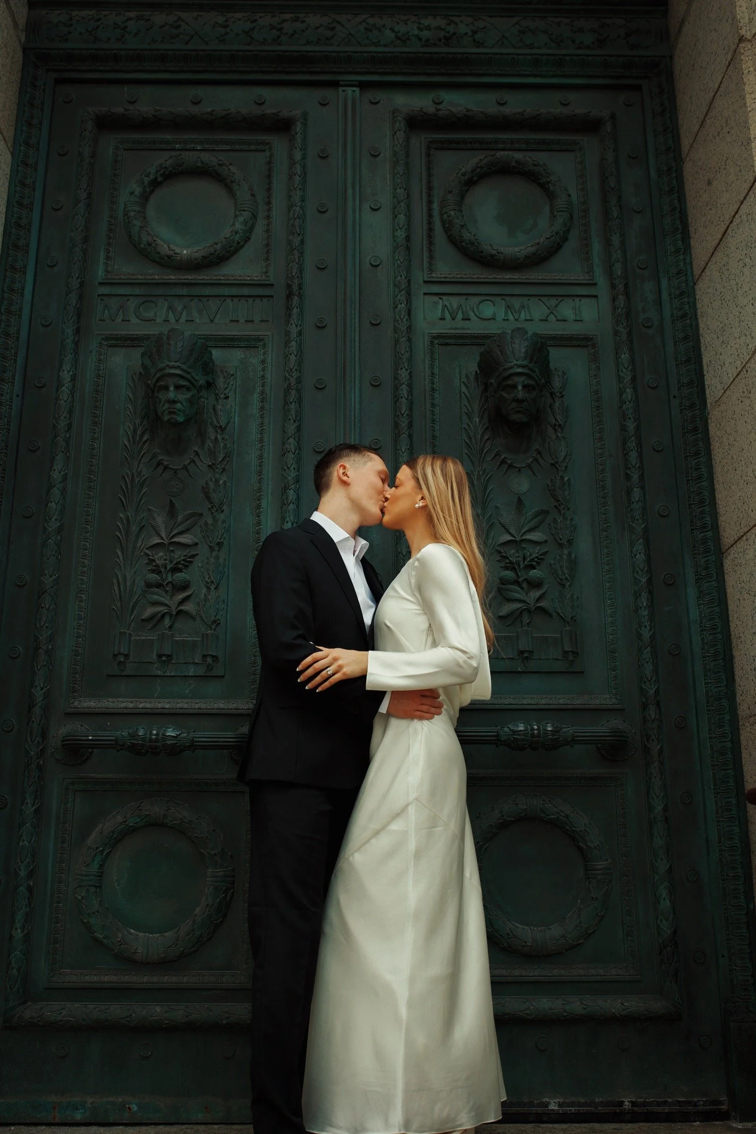 A couple in elegant attire sharing a kiss in front of a large ornate green bronze door with Roman numerals and carved faces.