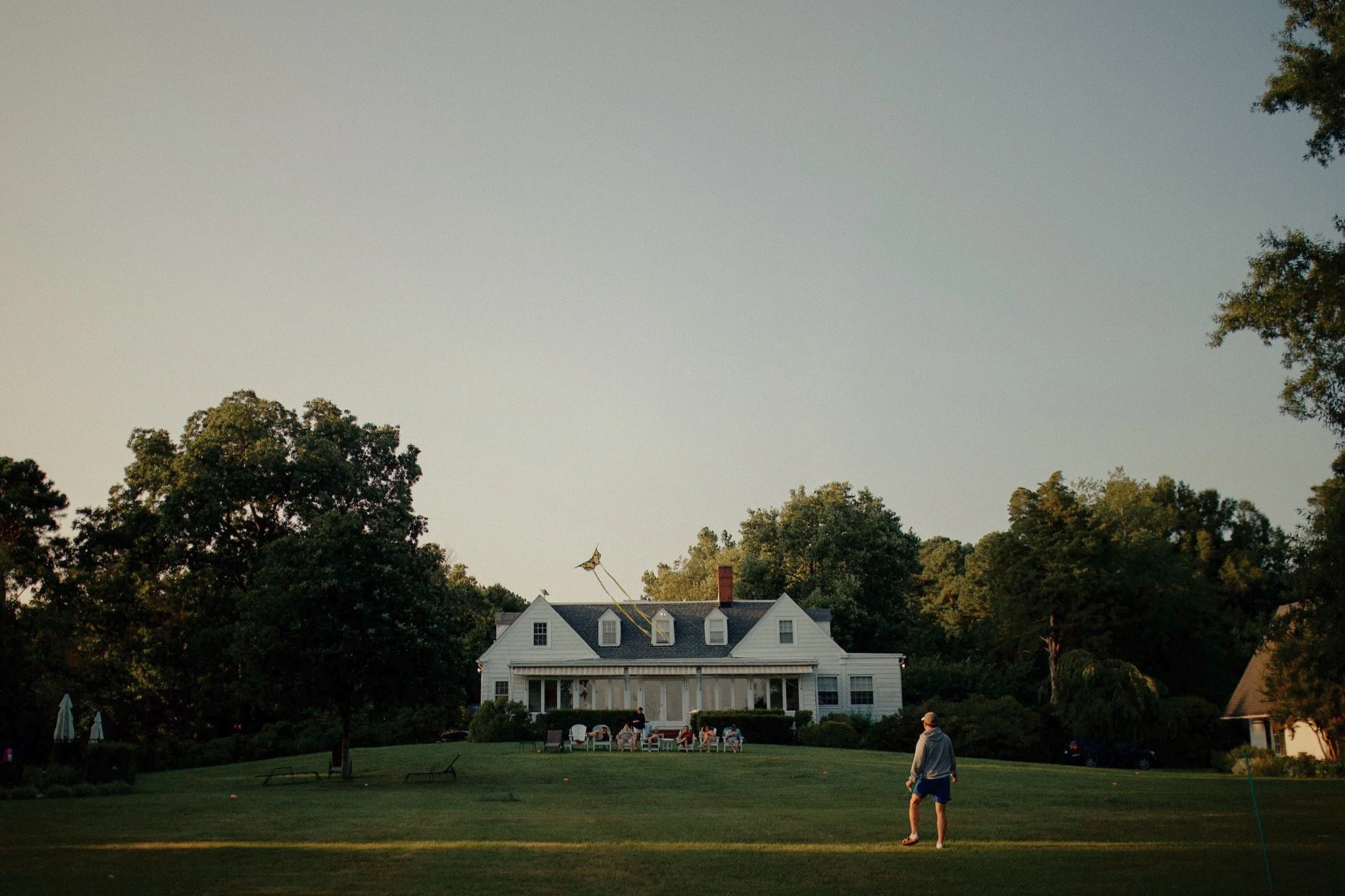 A man standing on grassy field in front of a white house with a large deck, surrounded by trees, during sunset.