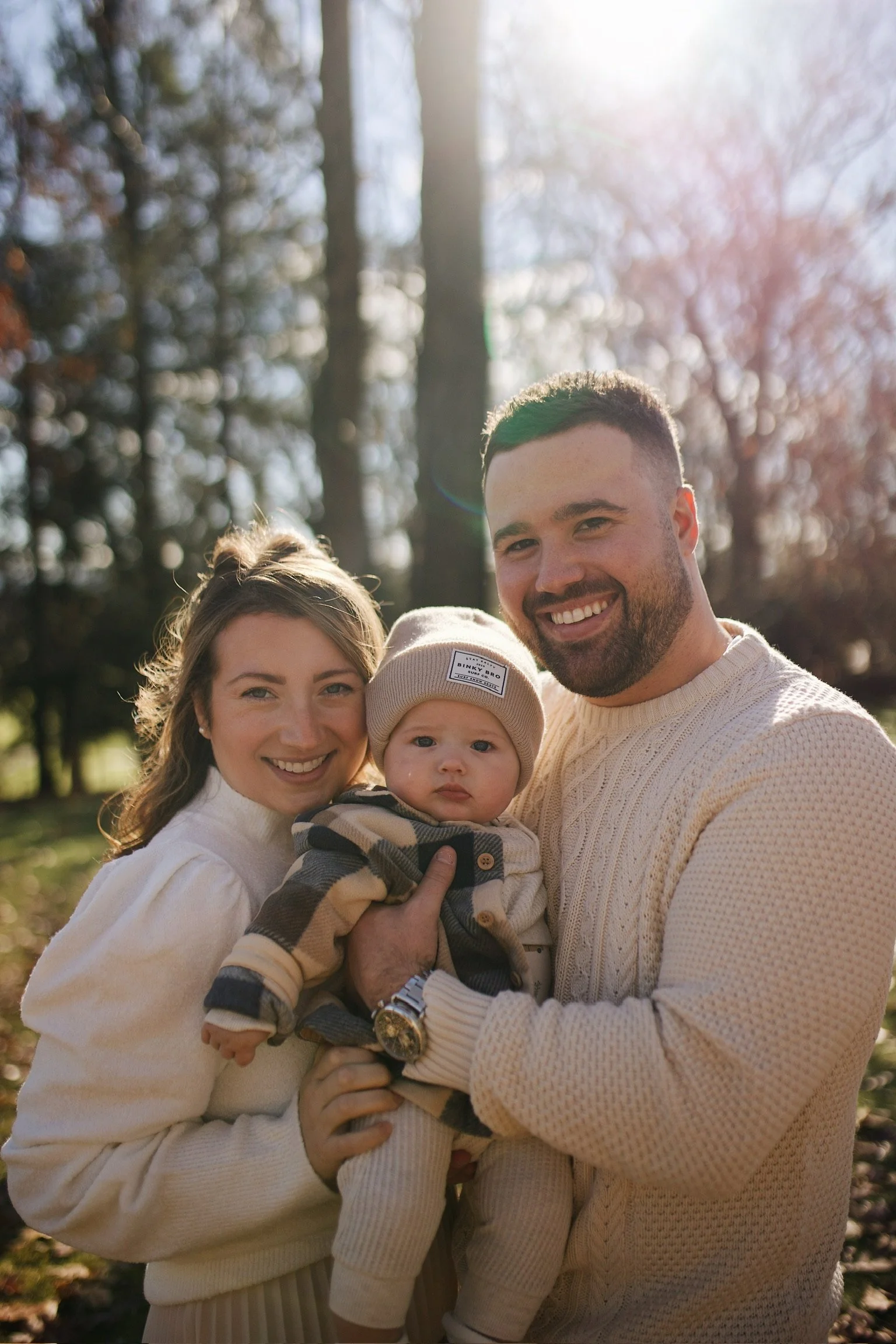A happy family of three outdoors in a park on a sunny day, with a woman, man, and baby girl, all smiling and dressed warmly for fall or winter.