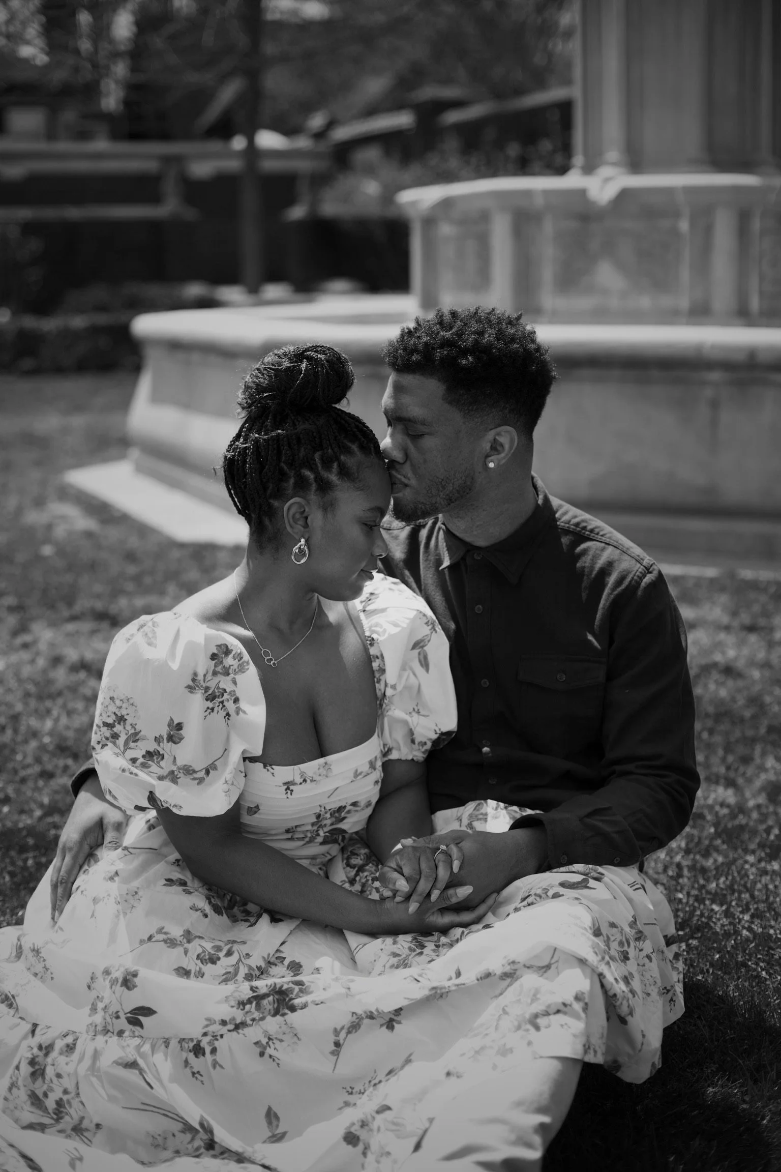 A black and white photo of a couple sitting on grass, holding hands, with the man kissing the woman’s forehead. The woman is wearing a floral dress and jewelry, and the man is in a dark button-up shirt. A fountain is in the background.
