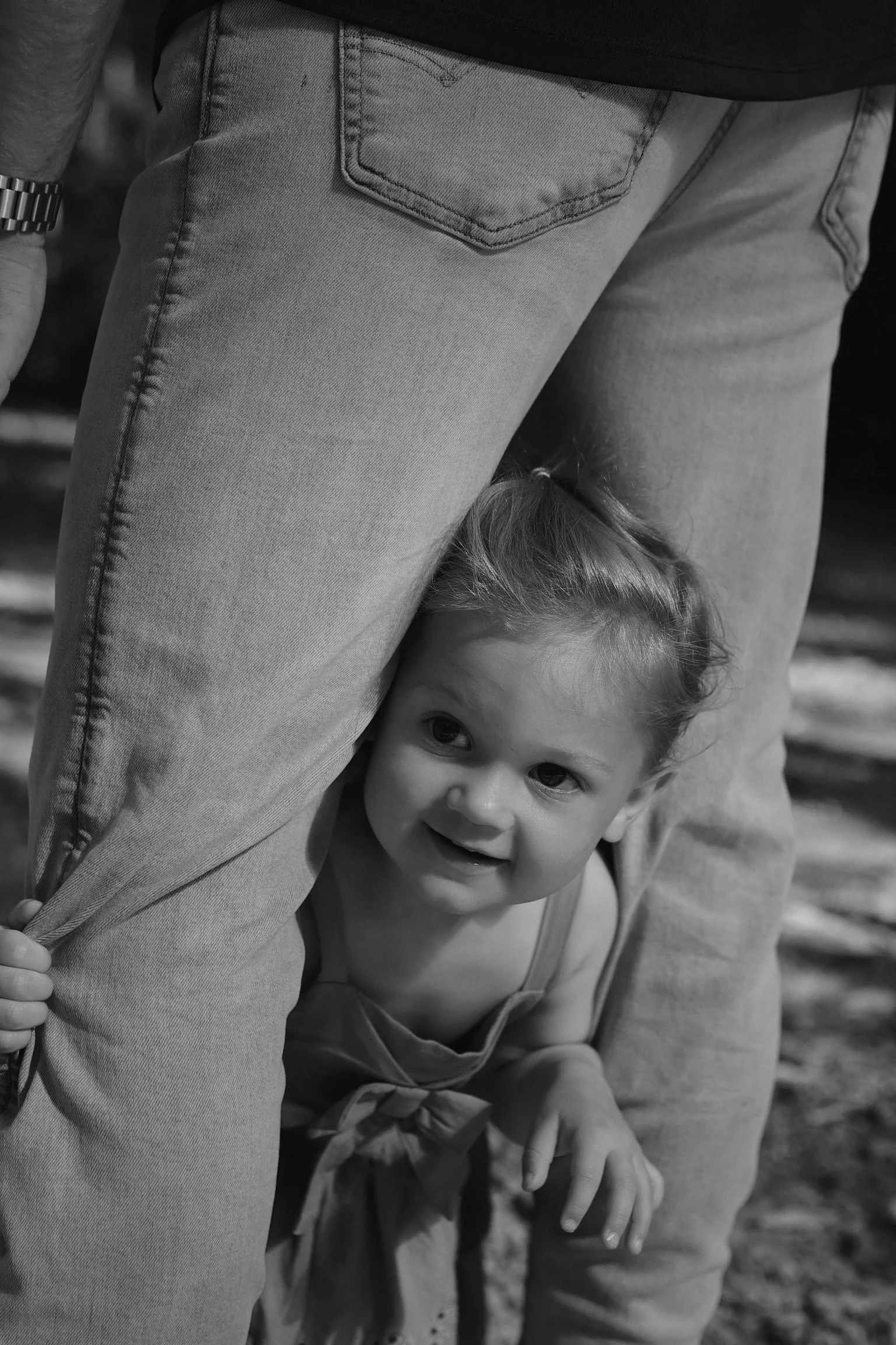 A young girl peeking out from behind a person's leg, smiling and looking at the camera, black and white photo.