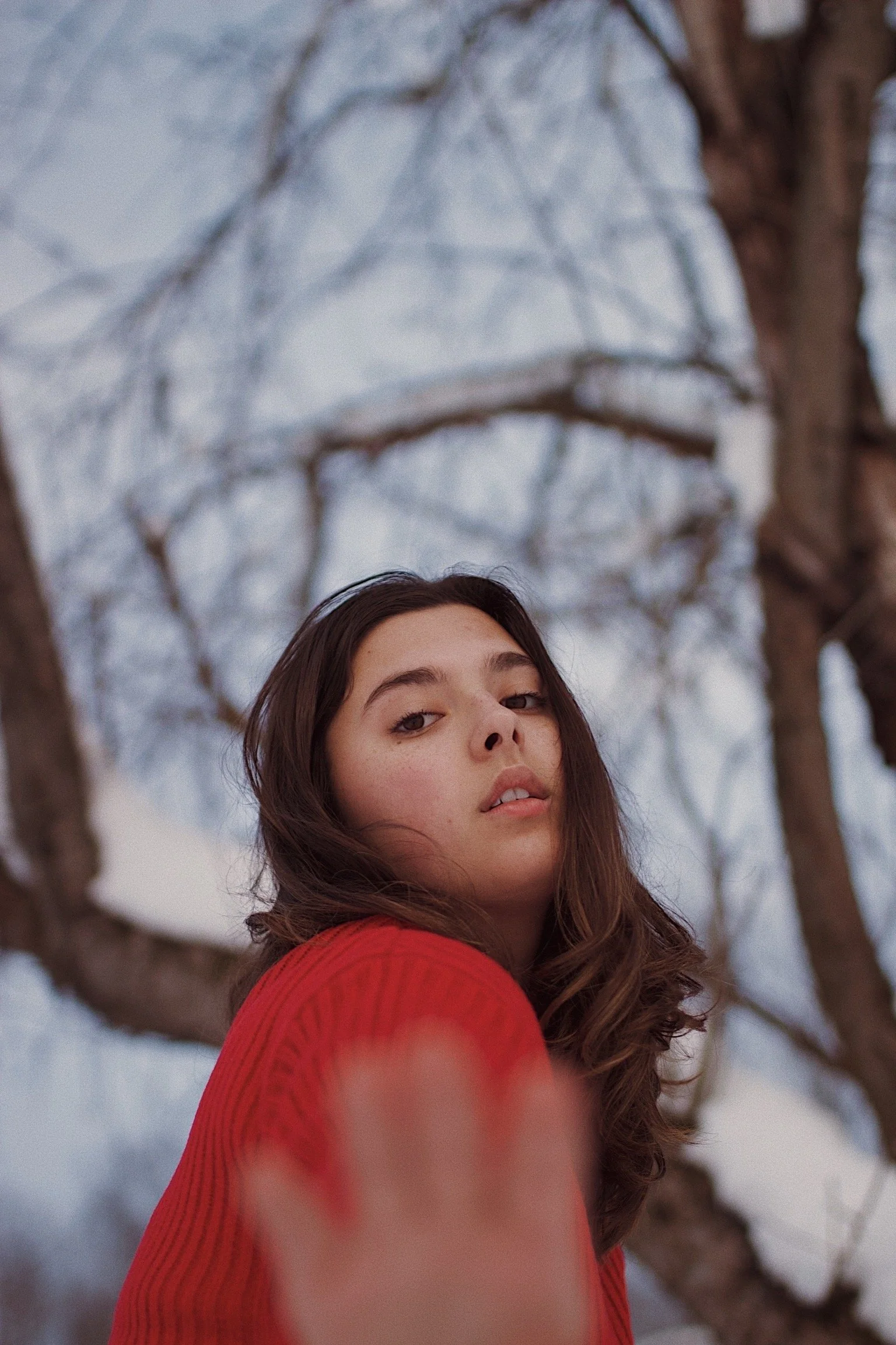 Young woman with long brown hair wearing a red sweater outdoors near a snow-covered tree