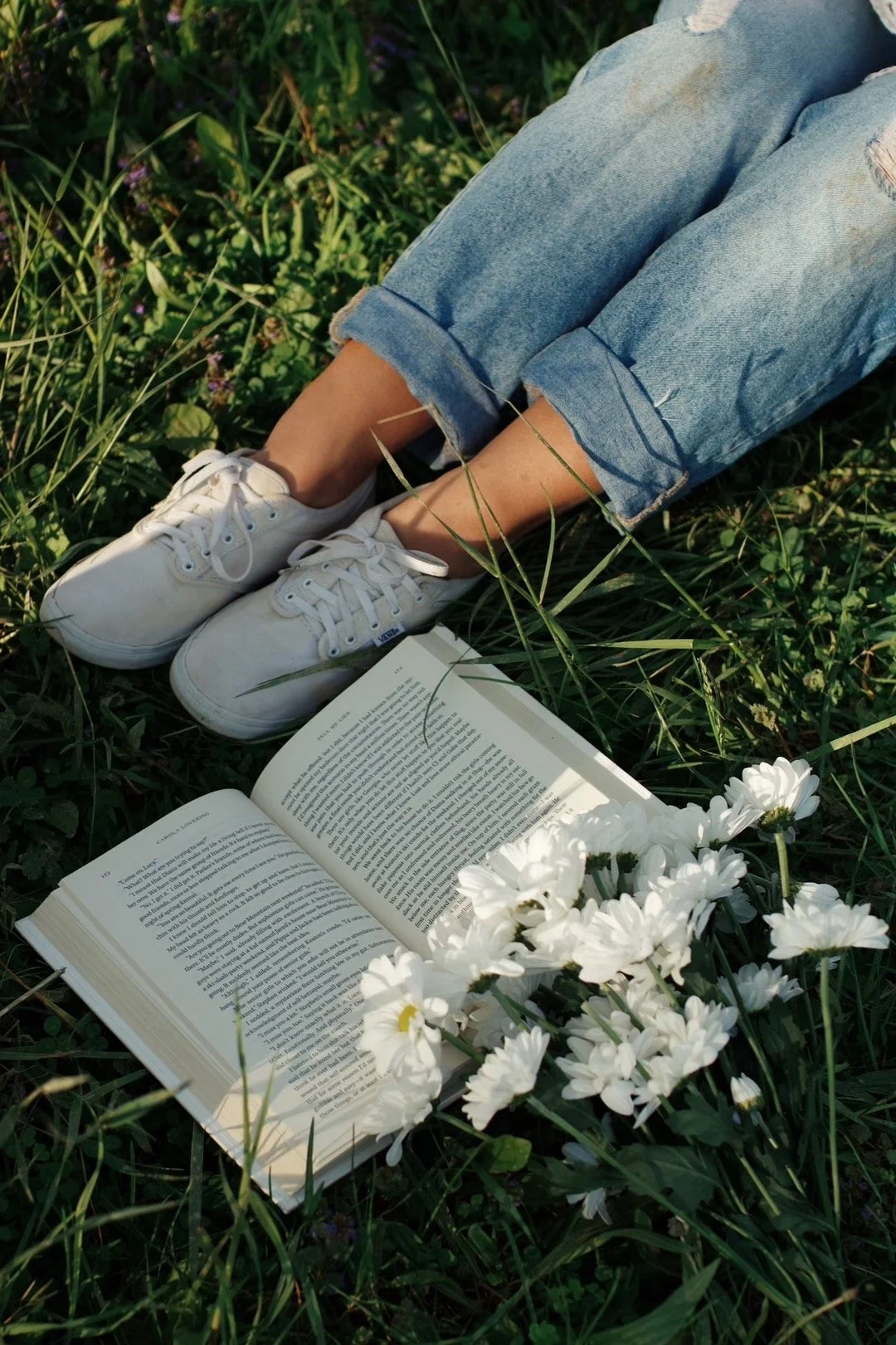 Person lying on grass wearing white sneakers and ripped jeans, with an open book and white daisies nearby.