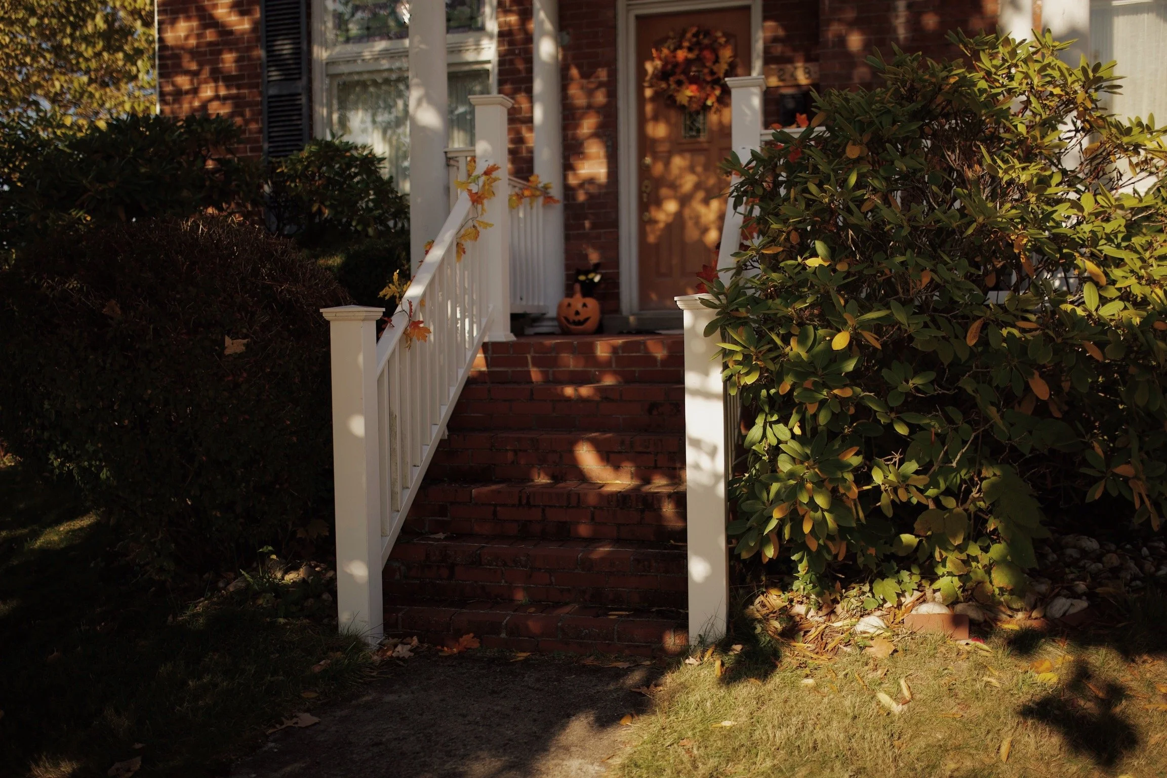 Front porch of a brick house with steps leading up, decorated for fall with seasonal foliage on the railing, a pumpkin decoration with a carved smile at the top of the stairs, and a wreath hanging on the door.