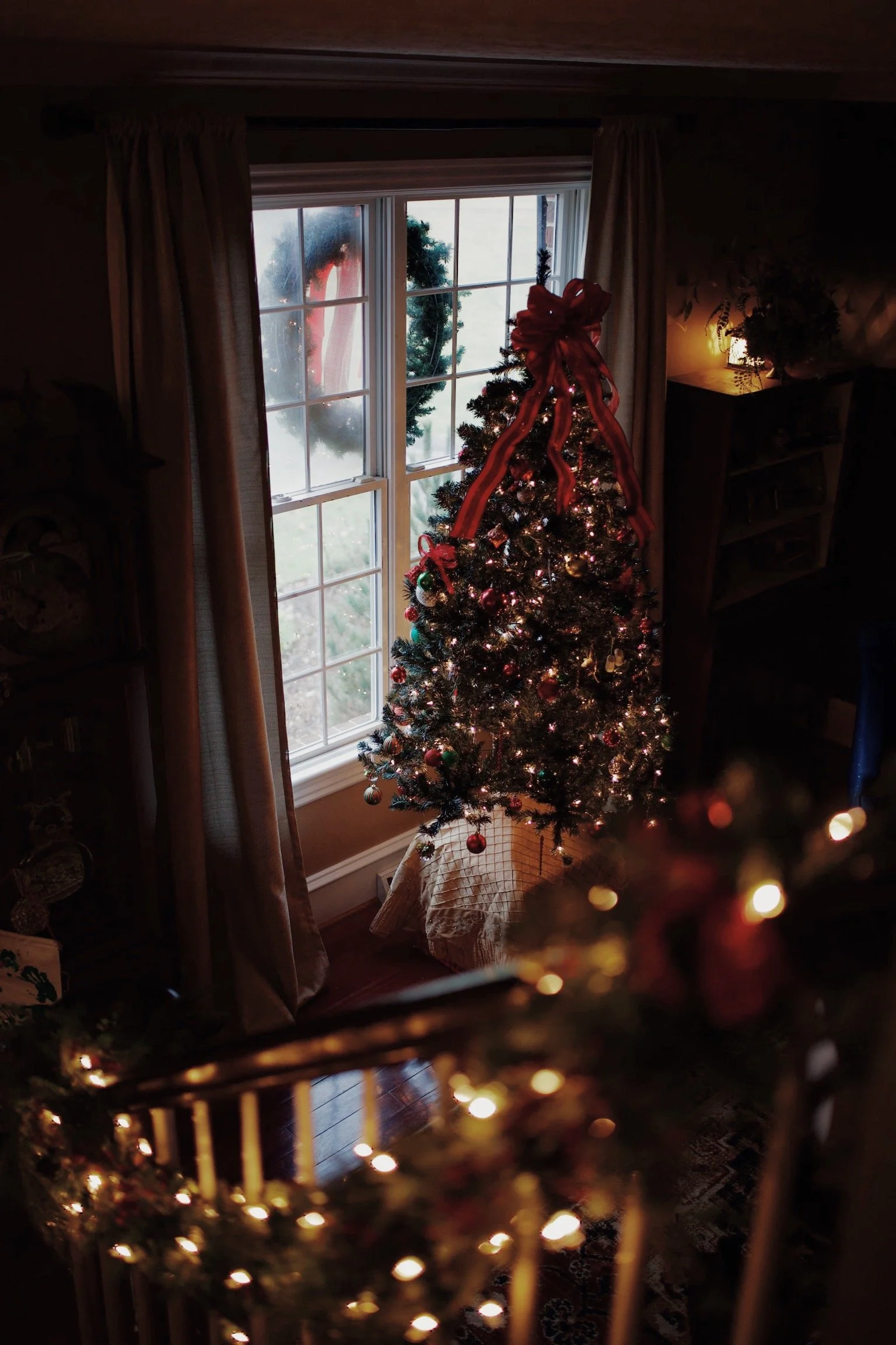 A decorated Christmas tree with lights, ornaments, and a red bow at the top, placed near a window with a wreath outside.