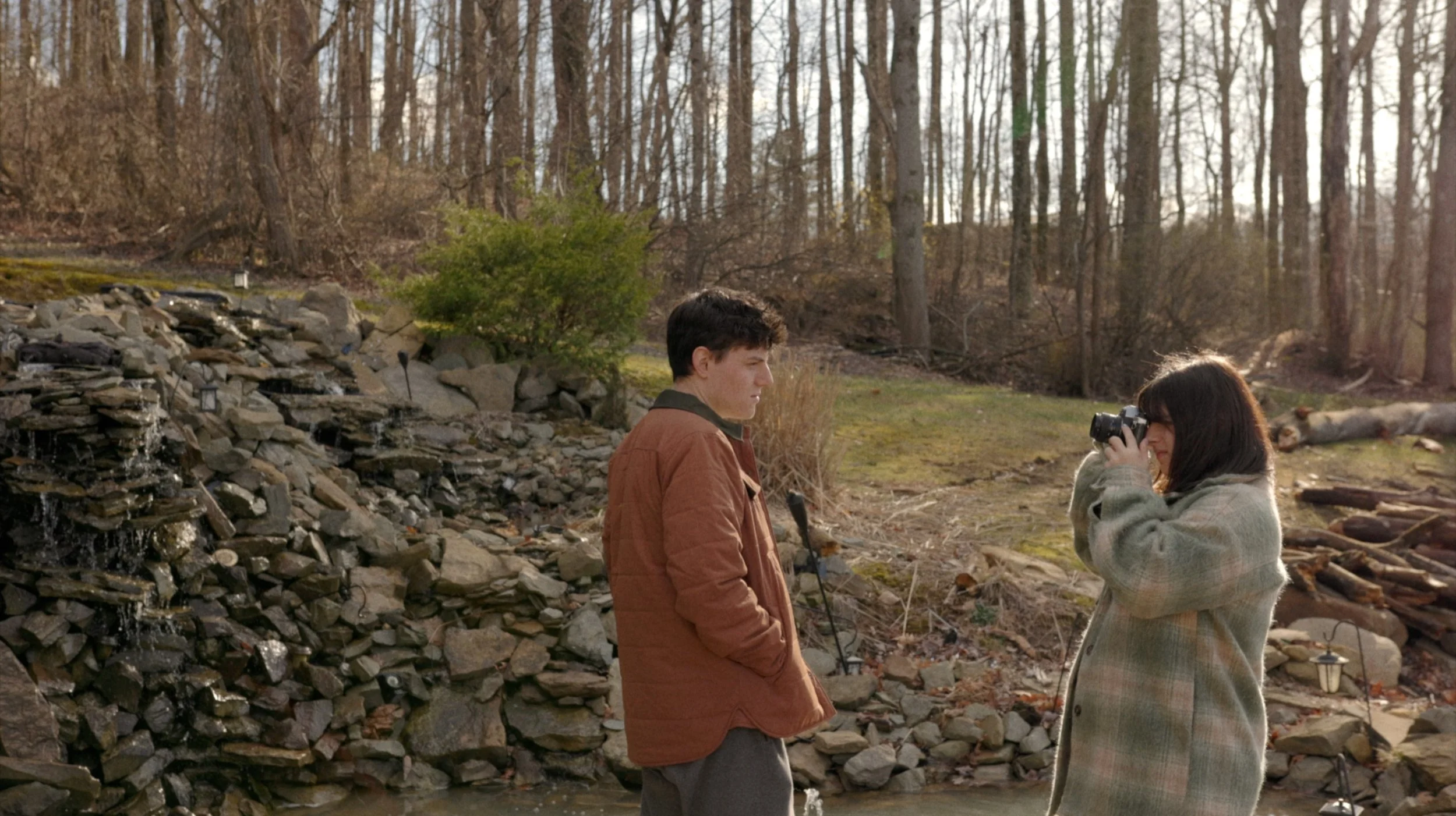 A young man standing near a rocky water feature in a wooded outdoor area, looking attentively at a woman taking a photo with a camera. The woman is wearing a checkered coat and hugging her camera to her face, with trees and sunlight in the background