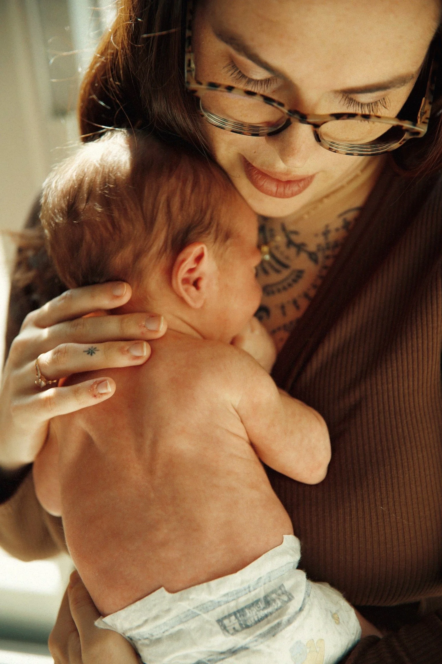 A woman with glasses gently holding and cuddling a newborn baby with reddish hair and a diaper, in a warm and intimate moment.