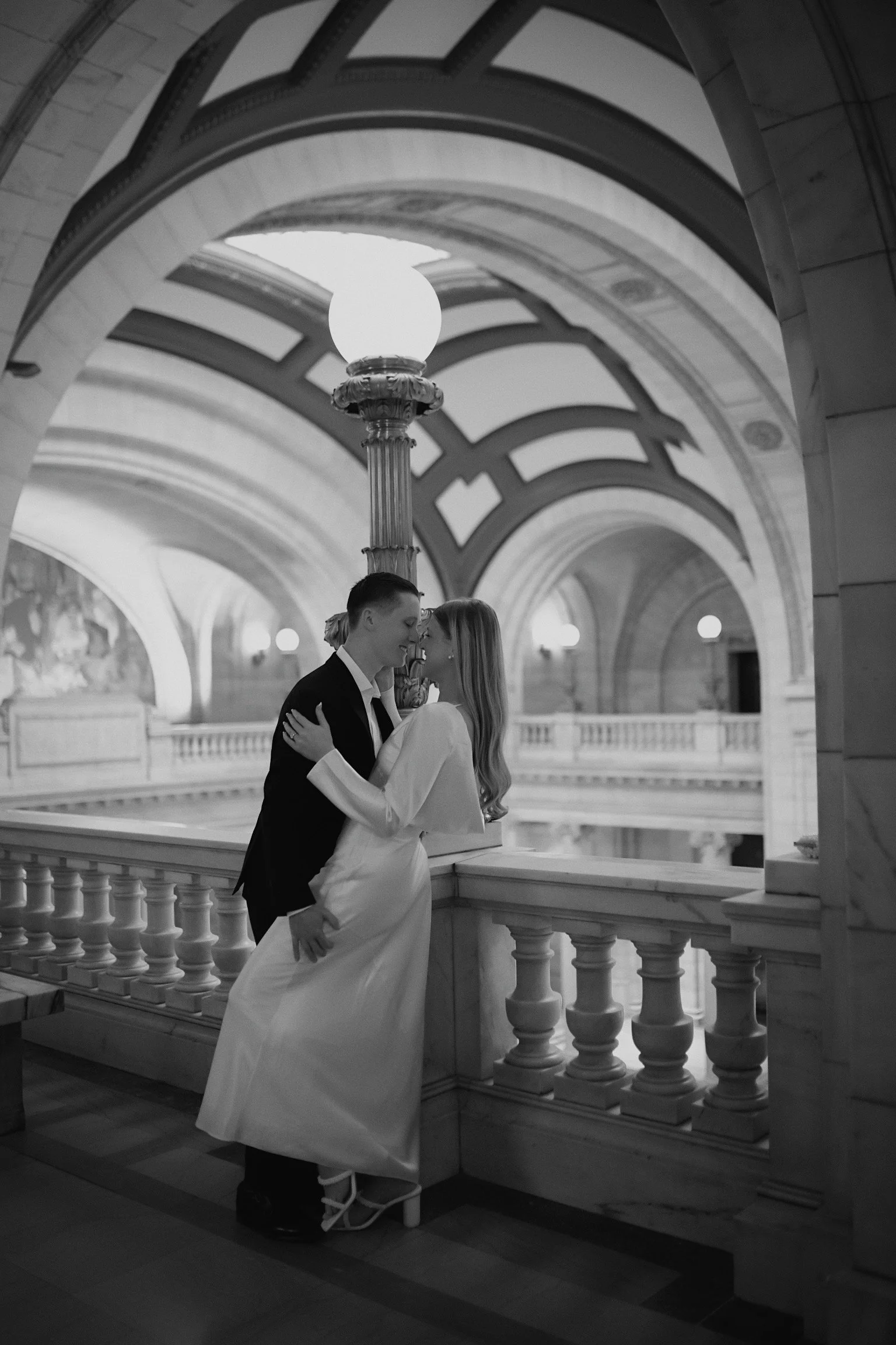 A couple in wedding attire sharing an intimate moment on a staircase inside a grand, historic building with ornate arches and a large decorative lamp post.