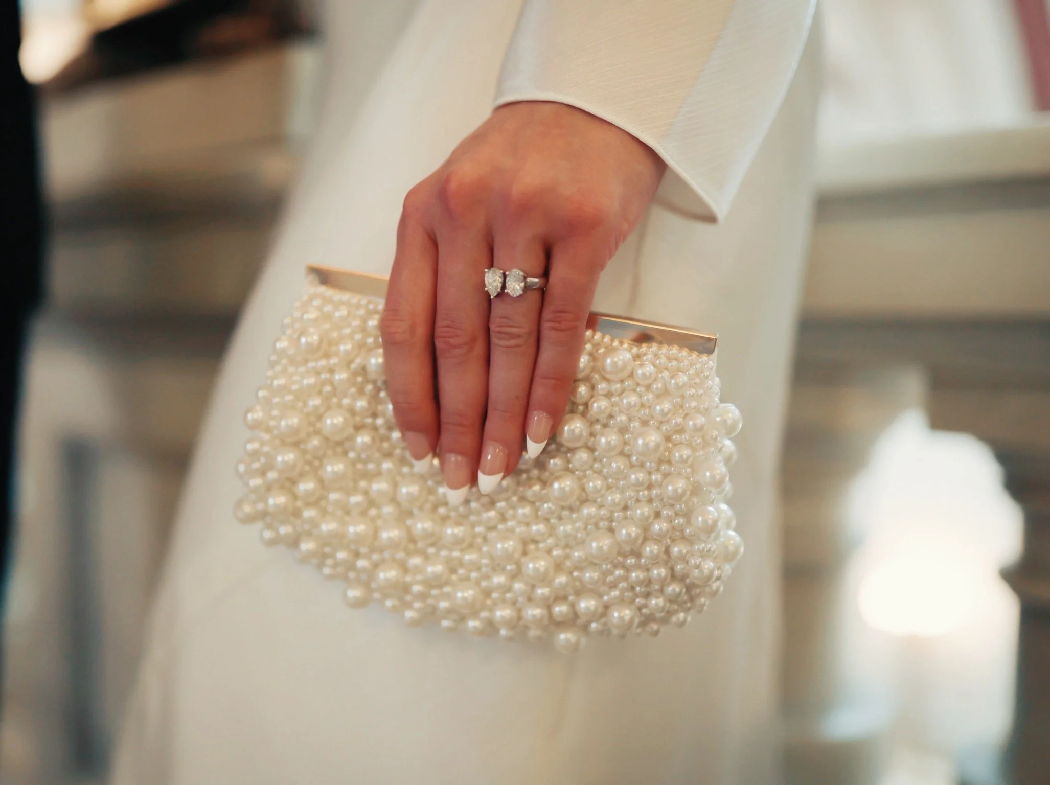 Close-up of a woman's hand holding a pearl-embellished clutch, wearing a diamond ring and a white sleeve.