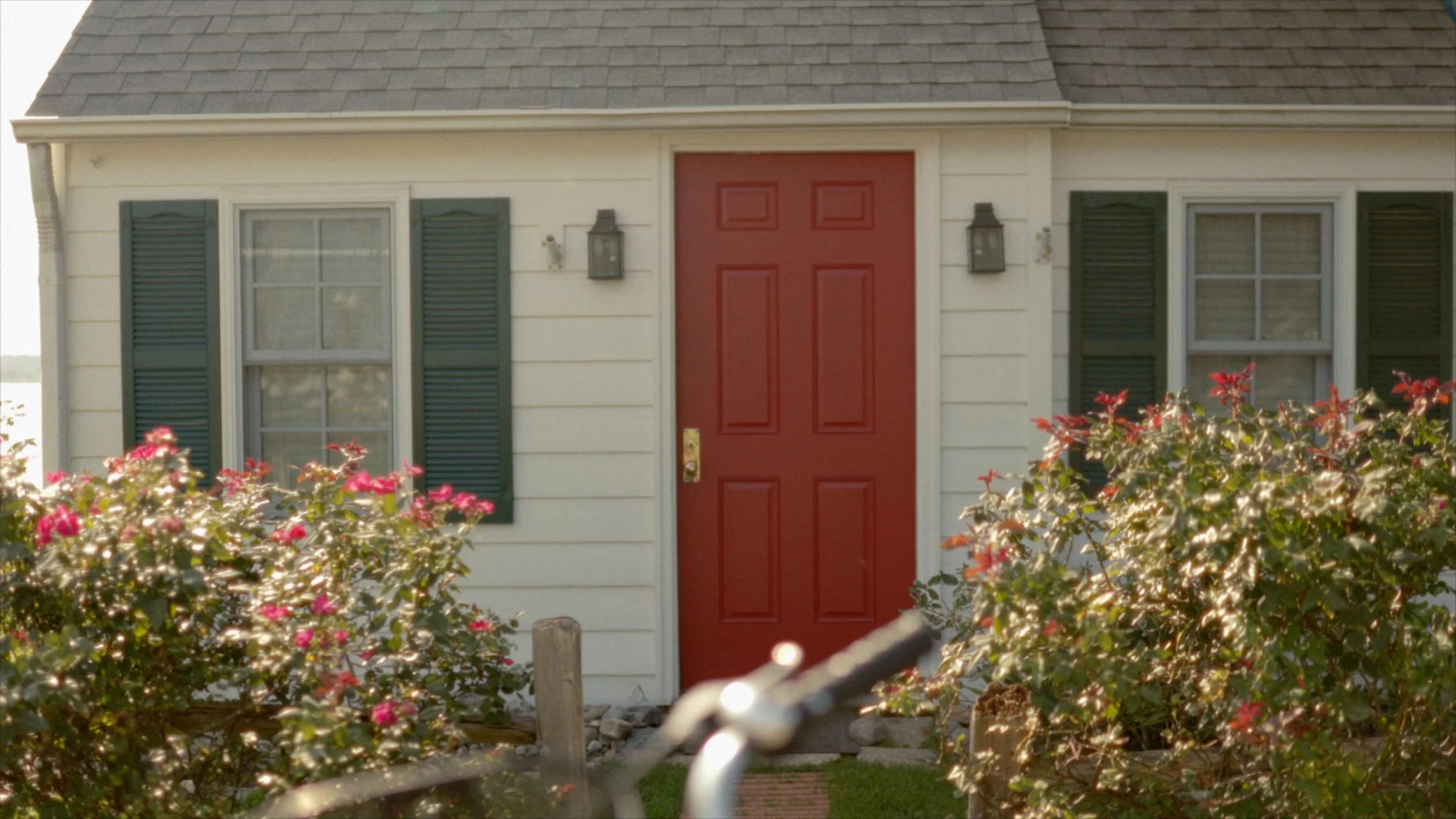 Front view of a house with a red door, white exterior walls, green shutters on the windows, and bushes with pink flowers in the foreground.