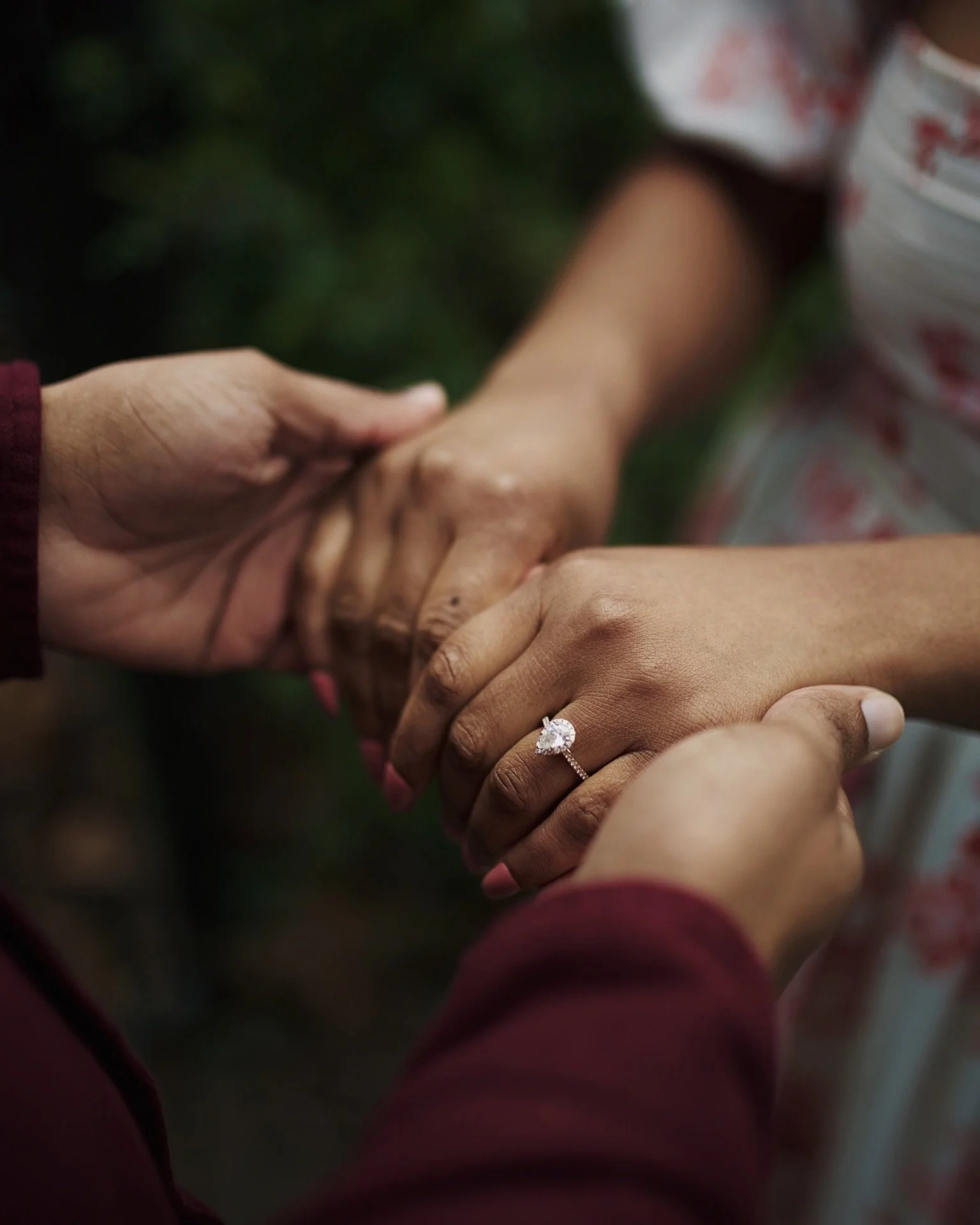 Close-up of two women holding hands, one wearing a large heart-shaped engagement ring with a diamond, with a blurred green background.