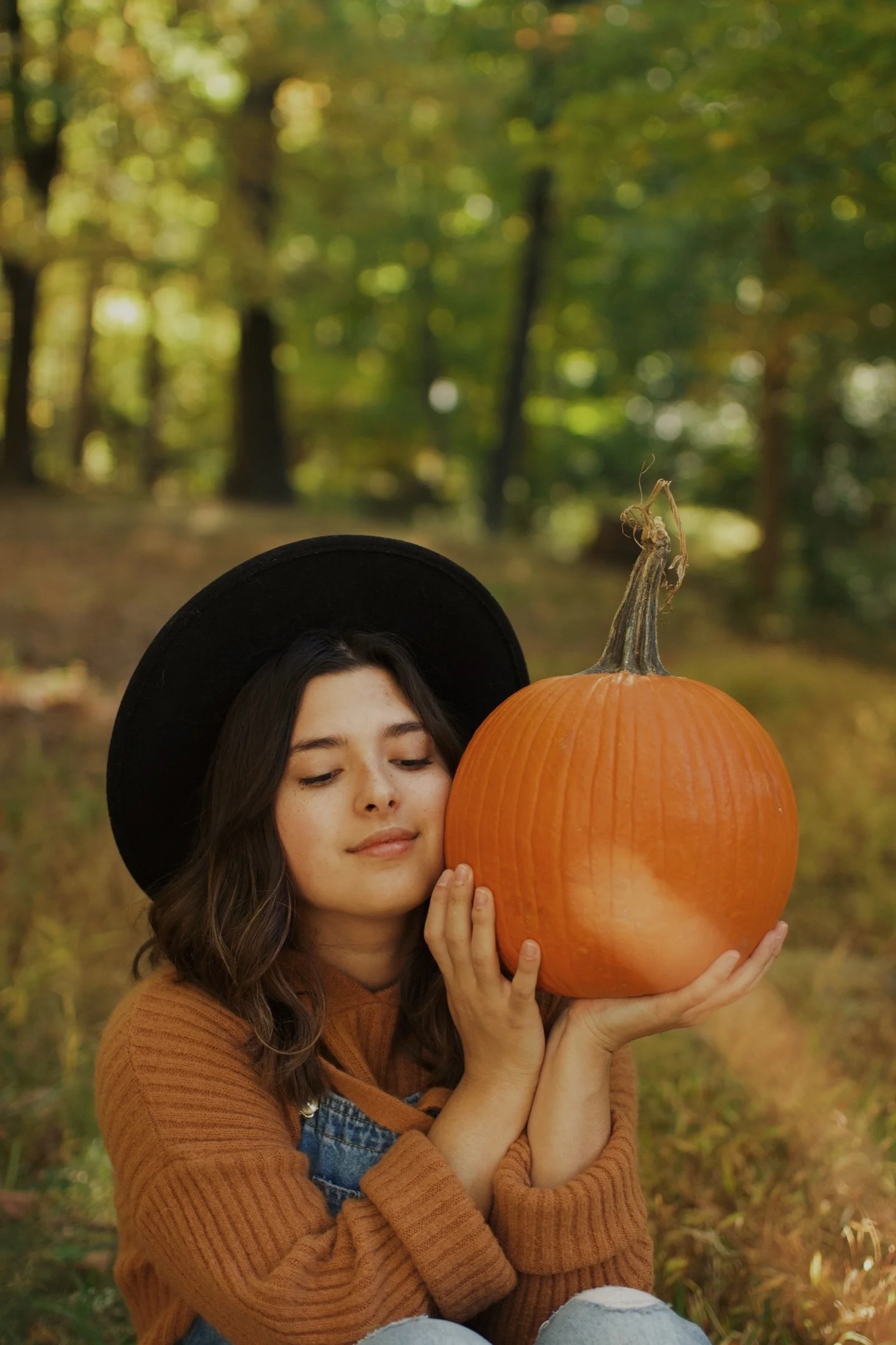 A young woman with a black hat, brown sweater, and overalls holding a large pumpkin in a forest with autumn leaves.