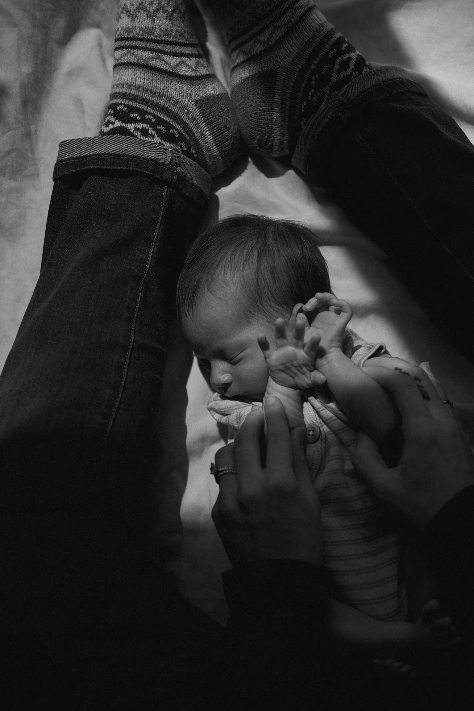 A baby sleeping peacefully on a bed with hands near face, surrounded by an adult’s legs and hands, in a black and white photo.
