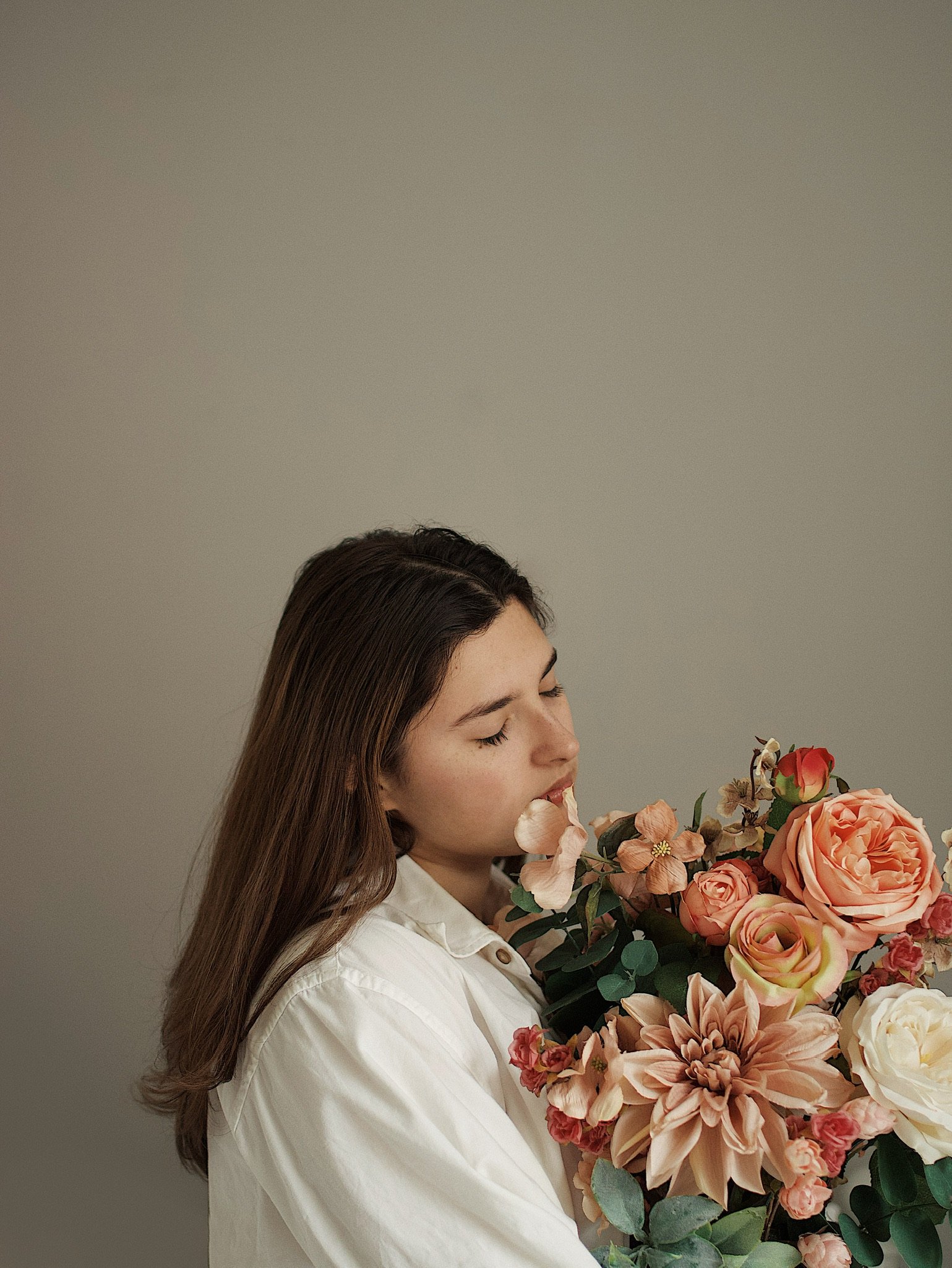 A woman with long brown hair wearing a white shirt holding a large bouquet of pink and white flowers, with some flowers close to her face.