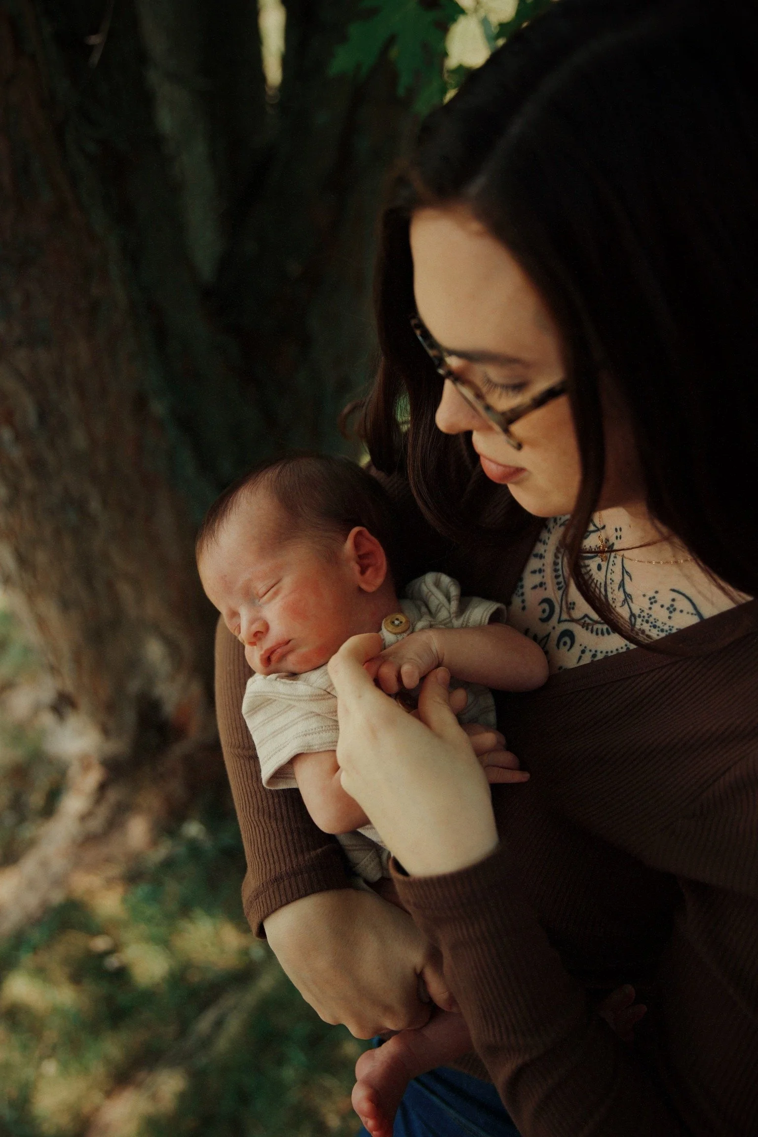 A woman holding a sleeping newborn baby close to her chest, gazing down affectionately, outdoors near a tree.