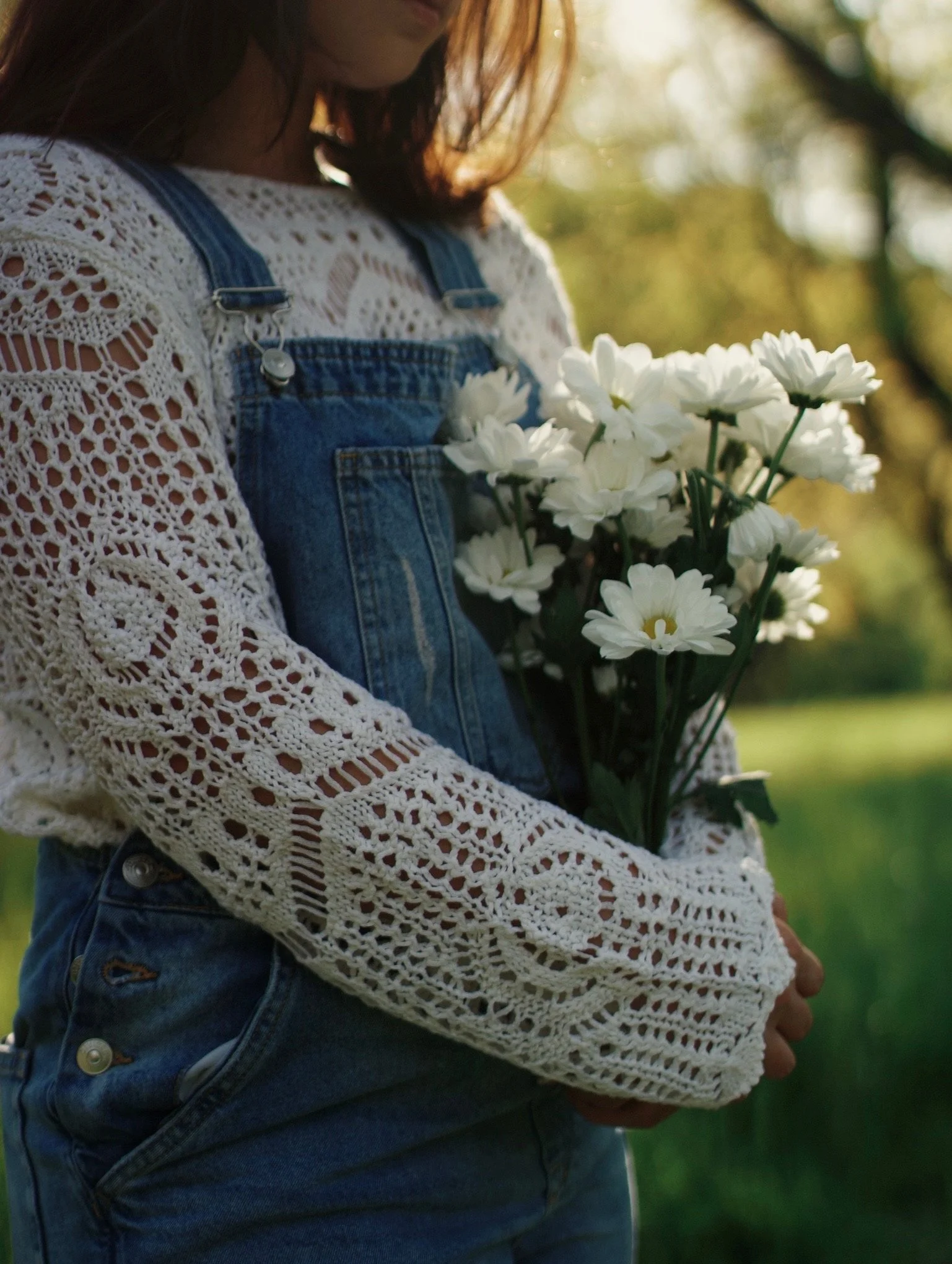 A woman in denim overalls and a white crochet top holding a bouquet of white daisies outdoors during daytime.