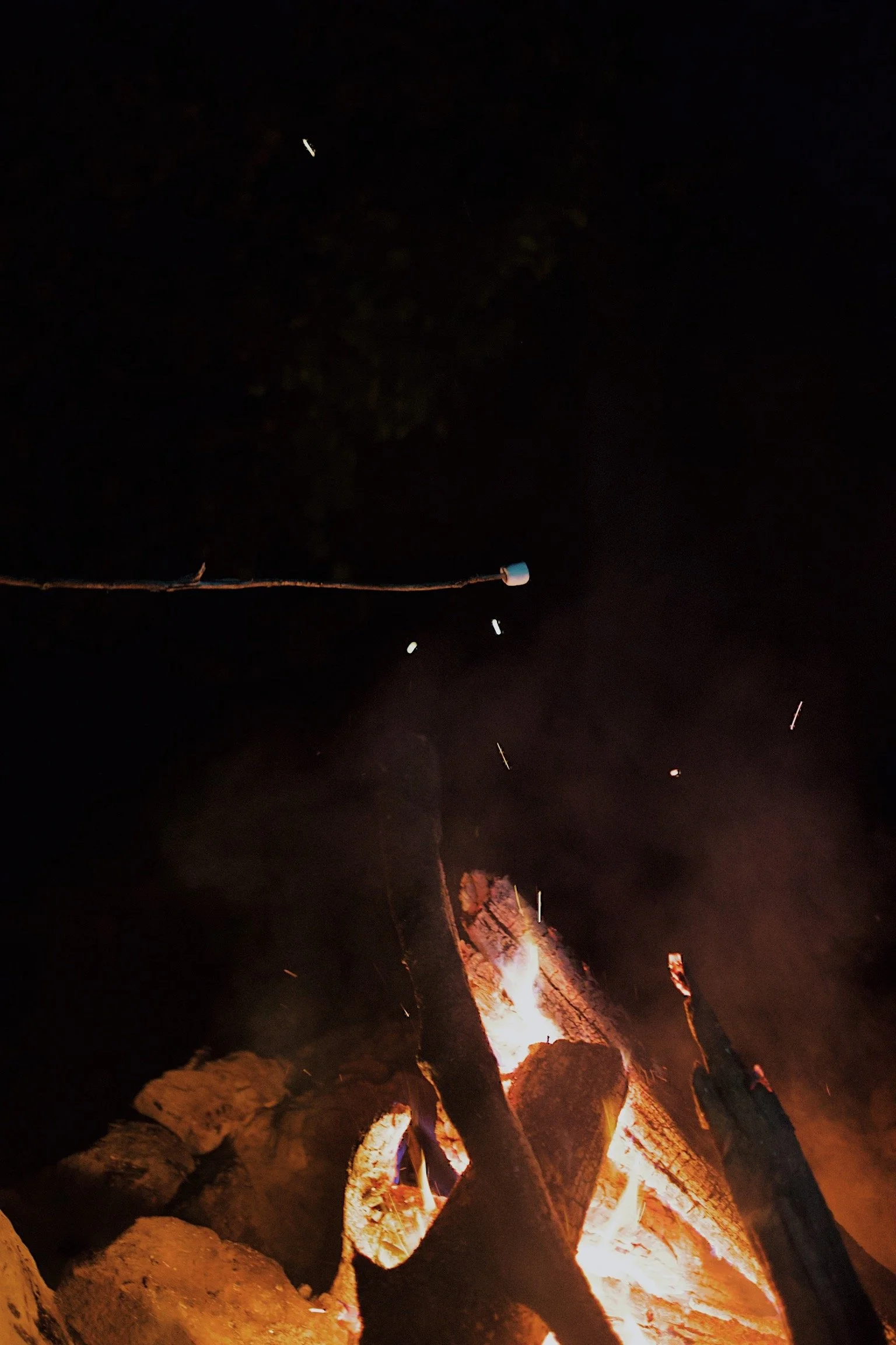Campfire with burning logs at night and a long stick with a marshmallow on the end.
