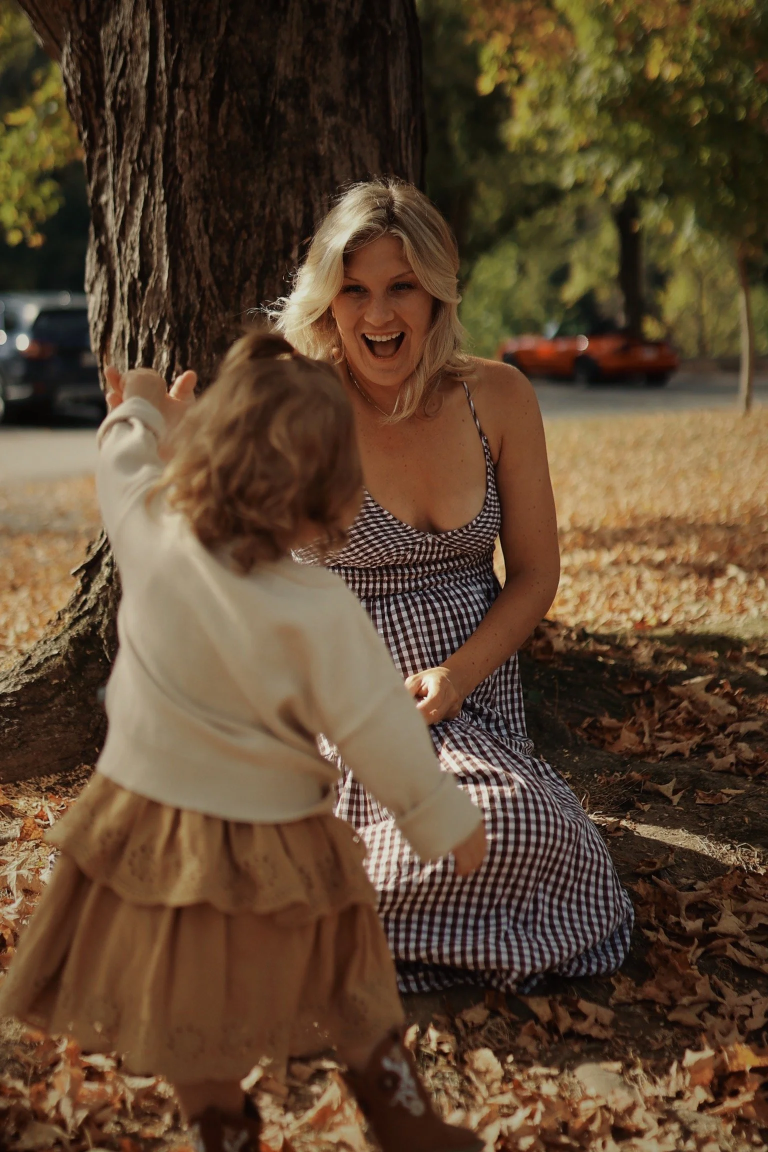 A woman with blonde hair in a checkered dress sitting under a tree, laughing and playing with a young child in a cream-colored jacket and a brown skirt, in a park covered with autumn leaves.