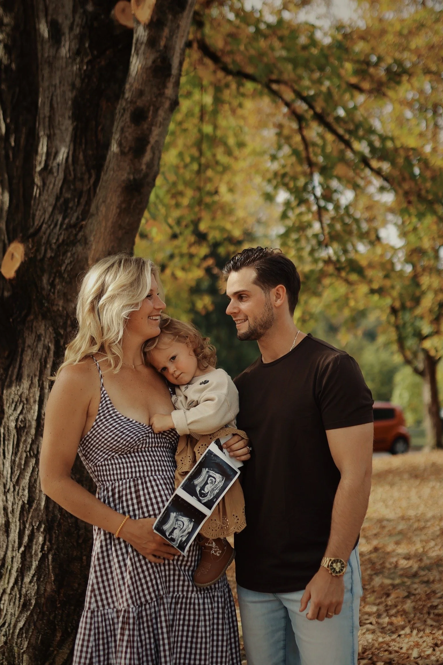 A family of three, including a pregnant woman, a man, and a young girl, standing outdoors near a large tree during autumn. The woman is holding ultrasound images, and the girl is resting her head on her mother's shoulder while the father looks on smi