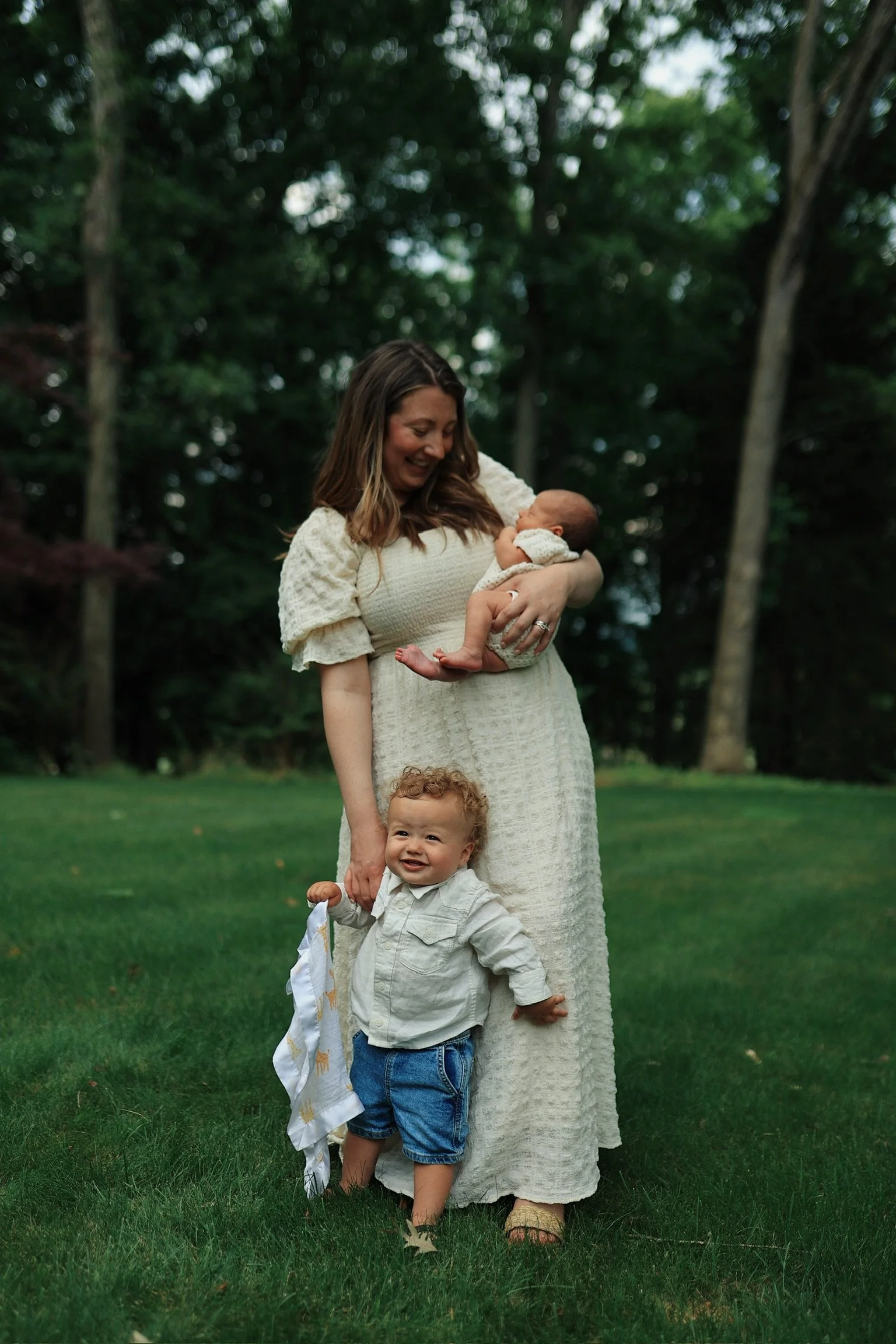 A woman holding a newborn baby in her arms and a toddler standing on the grass in front of her, smiling. They are outdoors in a park or garden with trees in the background.