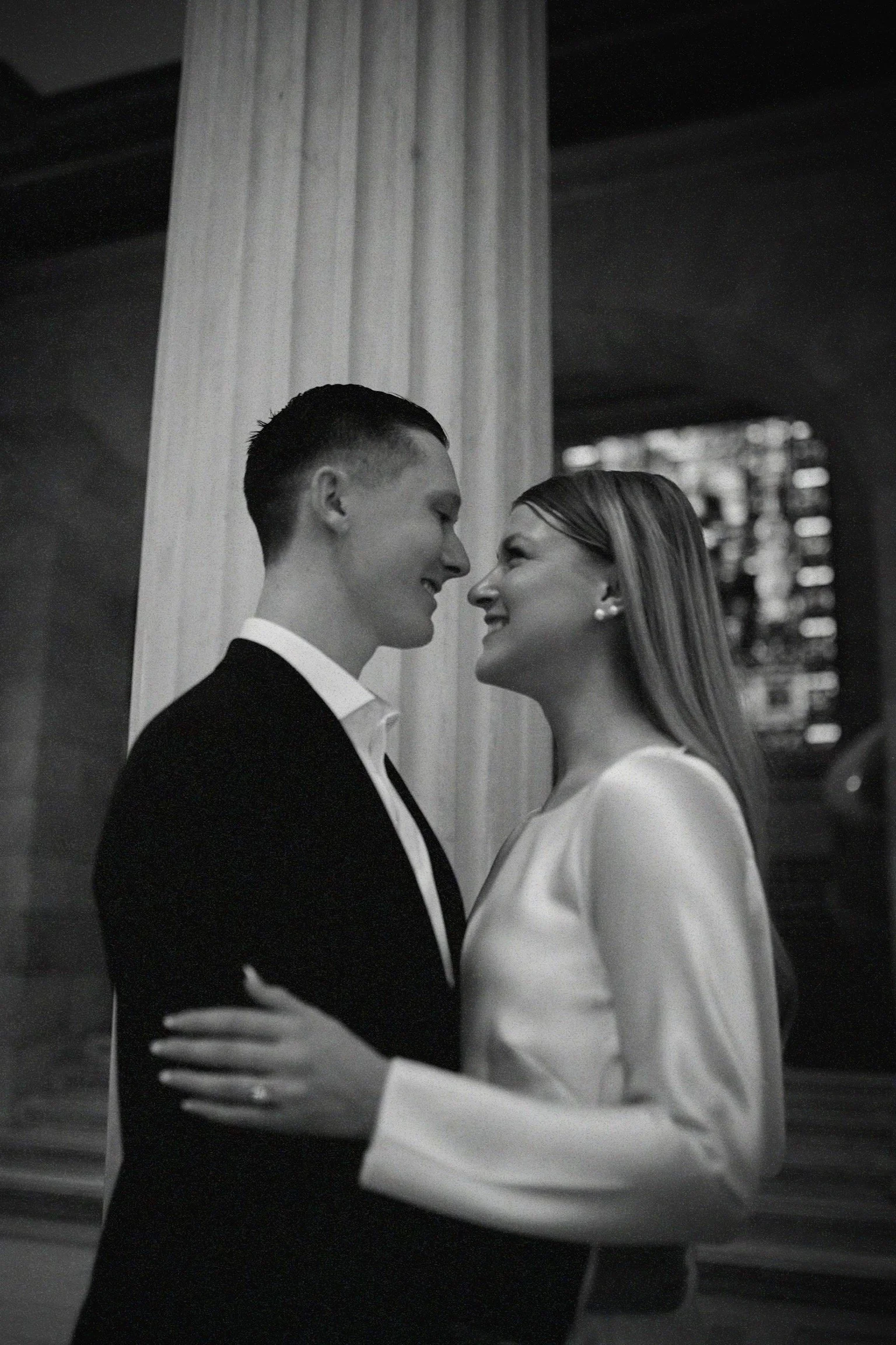 A black and white photo of a couple smiling and close to each other, embracing indoors with a column in the background.